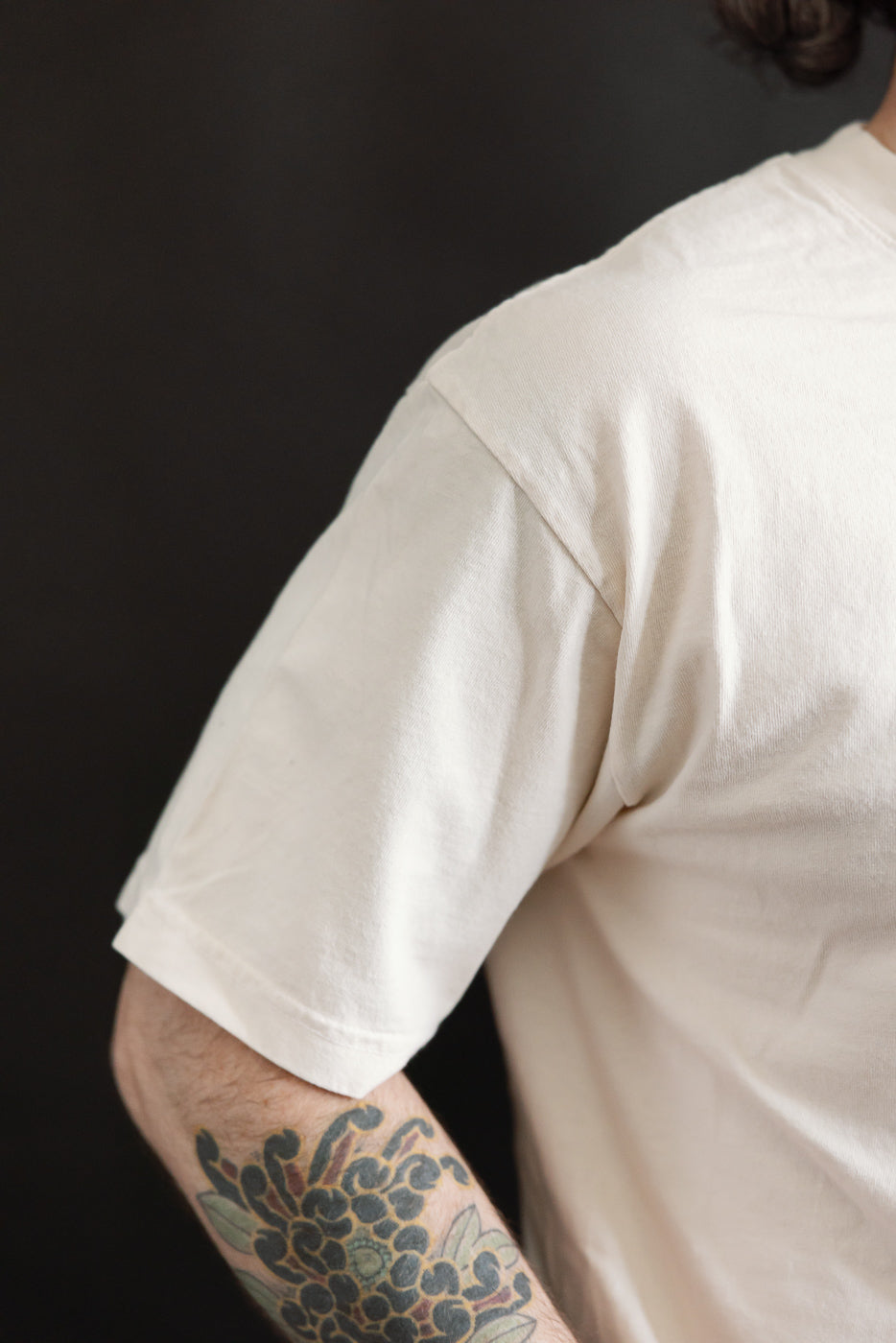 The 6.5oz - Dant Kingfisher T-Shirt - Ecru features a light-cream color with a vibrant Kingfisher bird design, showcasing a colorful tattoo on the forearm.