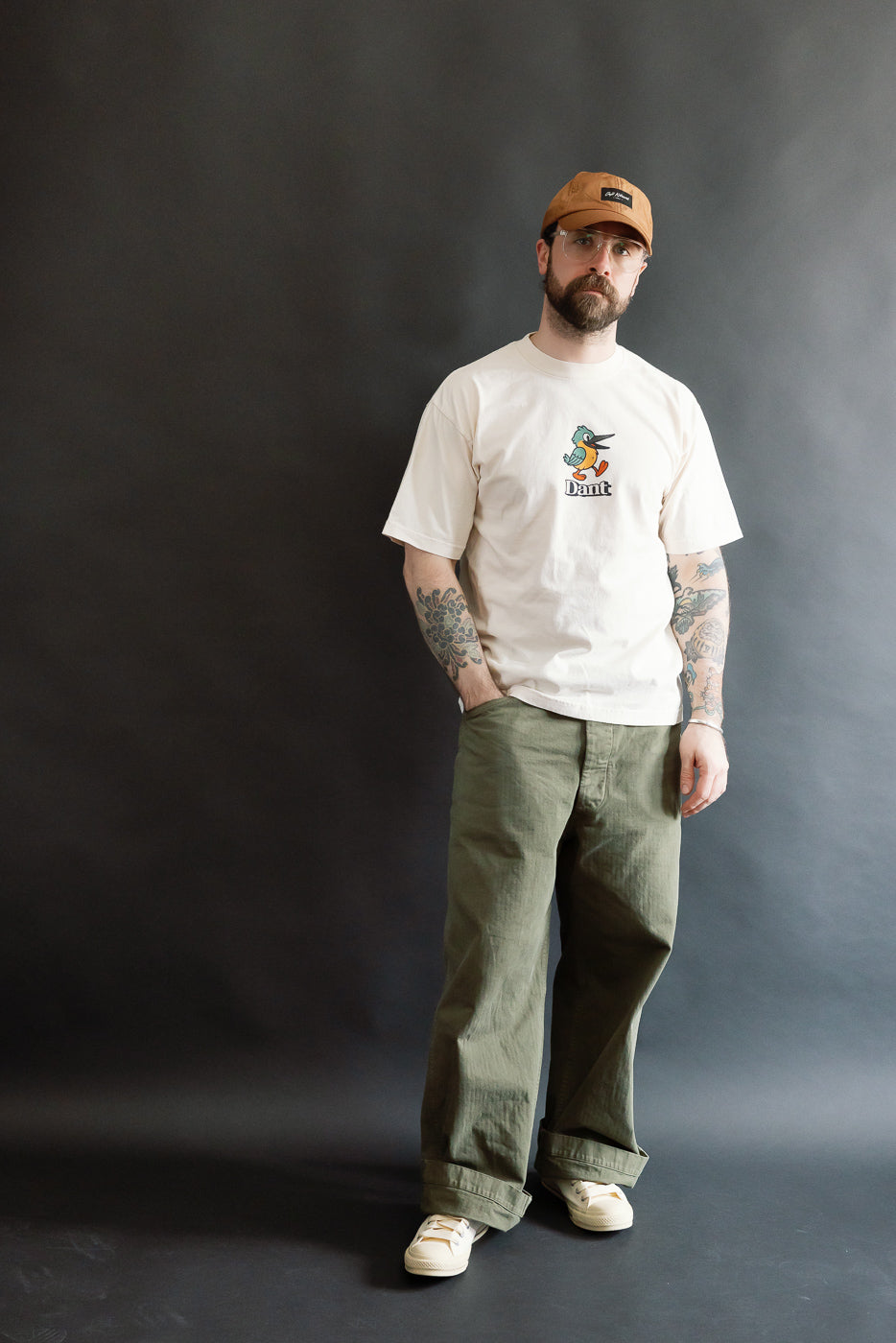 Man wearing the 6.5oz - Dant Kingfisher T-Shirt - Ecru with a cartoon duck print, styled with olive-green pants and off-white sneakers, showcasing Dant's relaxed, heritage-inspired fashion.