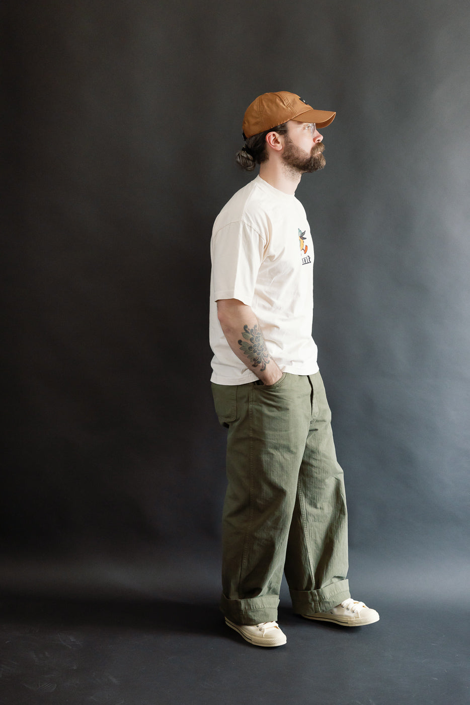 A studio portrait of a man wearing the 6.5oz - Dant Kingfisher T-Shirt - Ecru, showcasing its casual fit alongside olive trousers and cream sneakers, embodying Dant's heritage-inspired style.