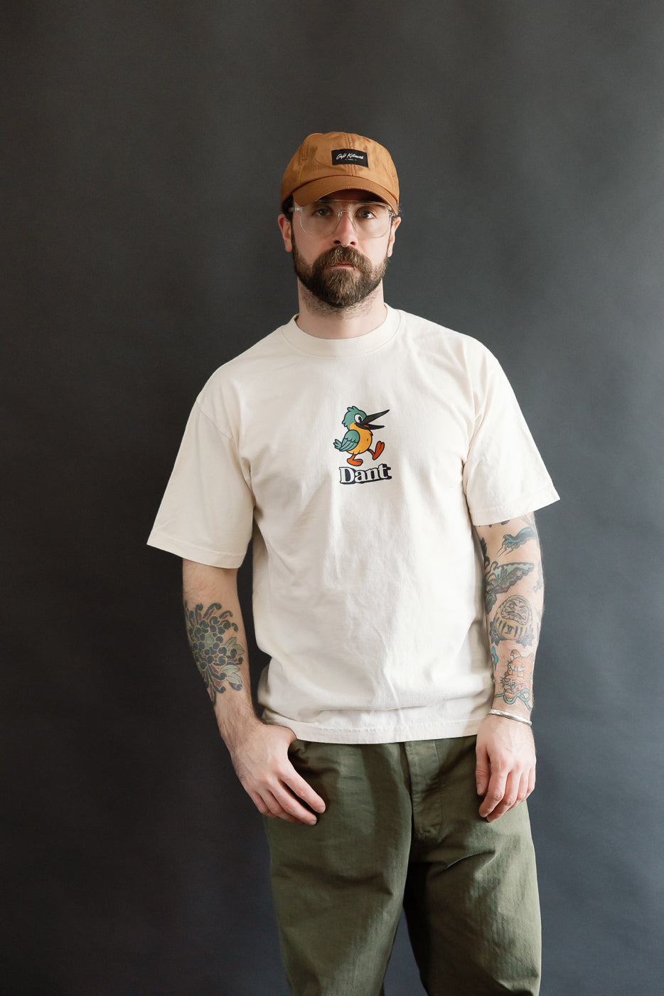 Model wearing the 6.5oz - Dant Kingfisher T-Shirt - Ecru, showcasing a cartoon bird design, tattoos visible on forearms, styled with olive-green pants and a light tan baseball cap.
