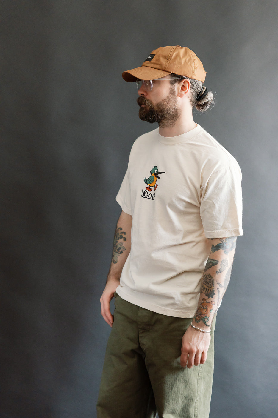 Bearded man wearing the 6.5oz Dant Kingfisher T-Shirt in Ecru, featuring a colorful cartoon duck logo. Styled with olive-green pants, showcasing Dant's blend of heritage and playful design.
