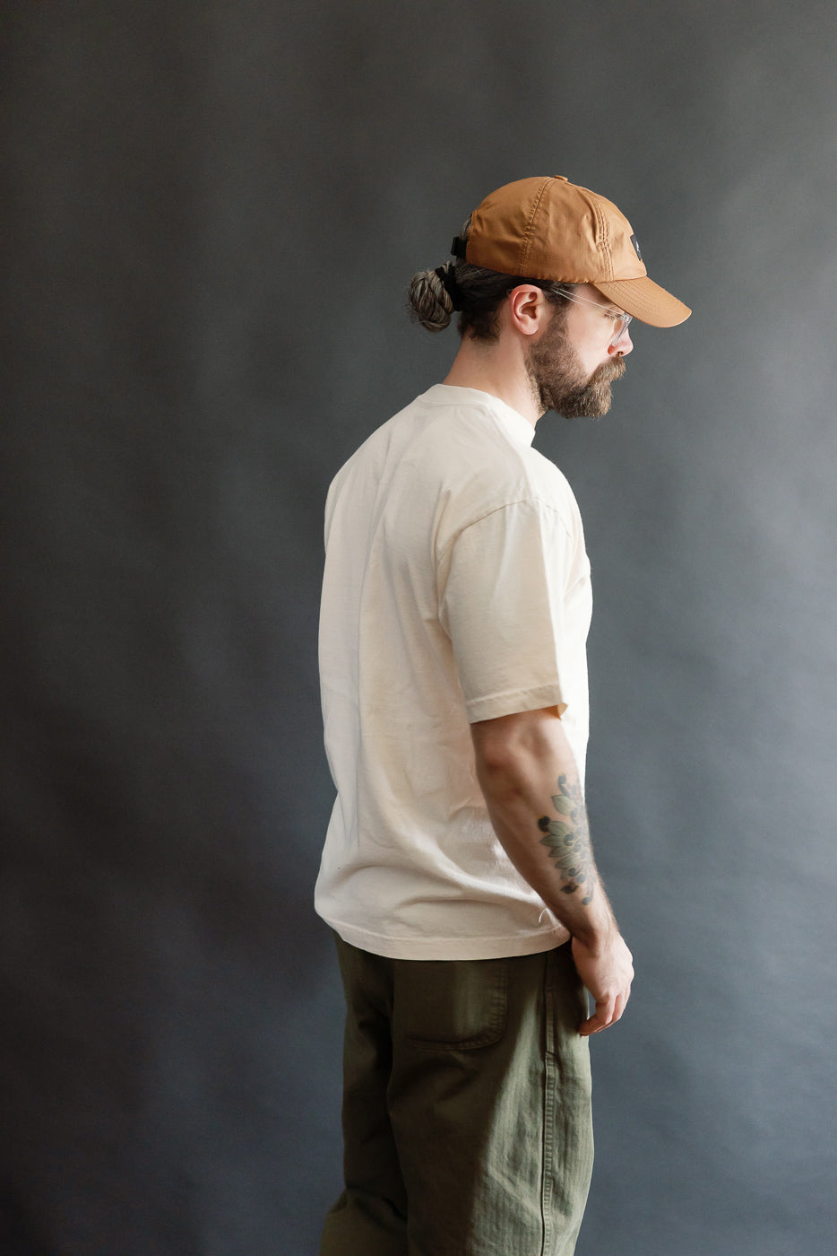 Young man in profile wearing a 6.5oz Dant Kingfisher T-Shirt - Ecru, showcasing a casual, heritage-inspired look with olive-green trousers and a brown baseball cap, emphasizing the shirt's oversized, modern fit.
