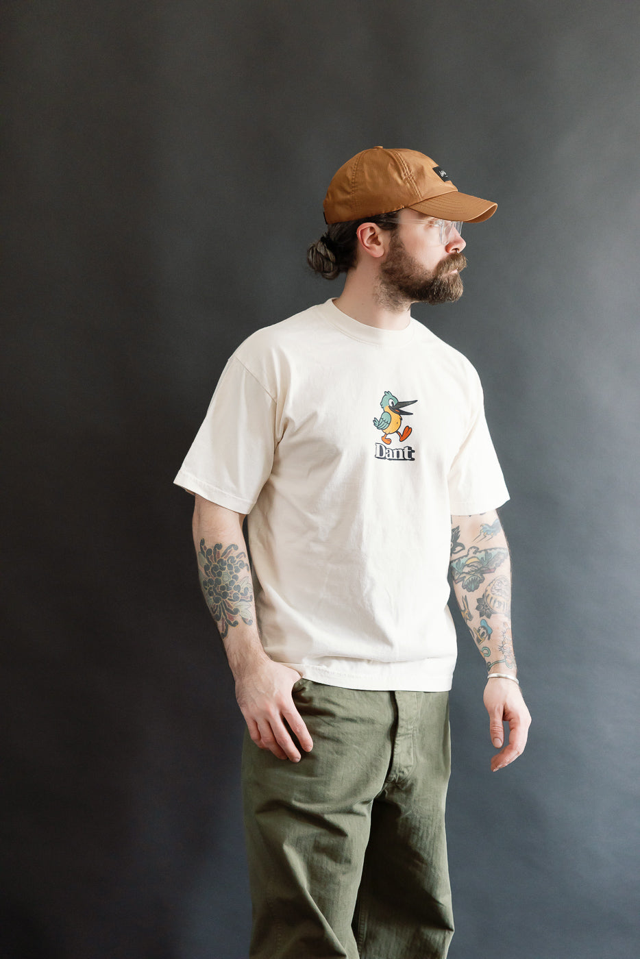 Model wearing the 6.5oz - Dant Kingfisher T-Shirt - Ecru, featuring a small cartoon bird print, styled with olive-green pants and a brown cap against a gray backdrop.