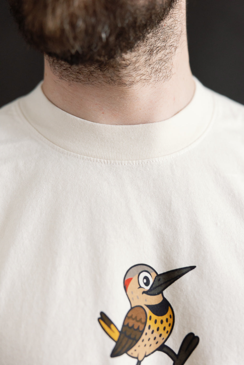 6.5oz - Dant Northern Flicker T-Shirt - Ecru features a cartoon woodpecker on a light-colored shirt, reflecting a playful design inspired by birdwatching, merging nature with Dant's heritage-inspired style.