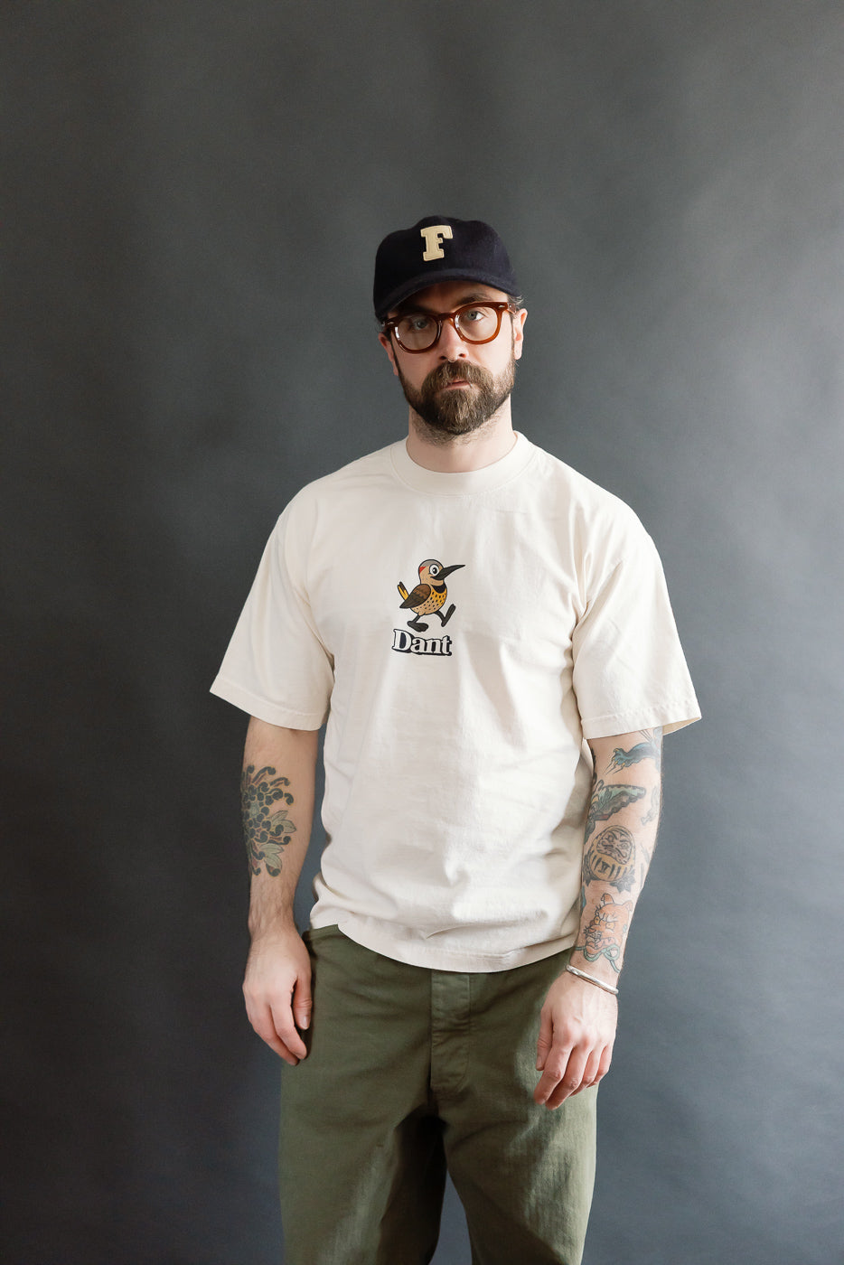 Man wearing the 6.5oz - Dant Northern Flicker T-Shirt - Ecru, featuring a cartoon bird and Dant text, paired with olive-green trousers and a black cap, showcasing a casual, modern fit.