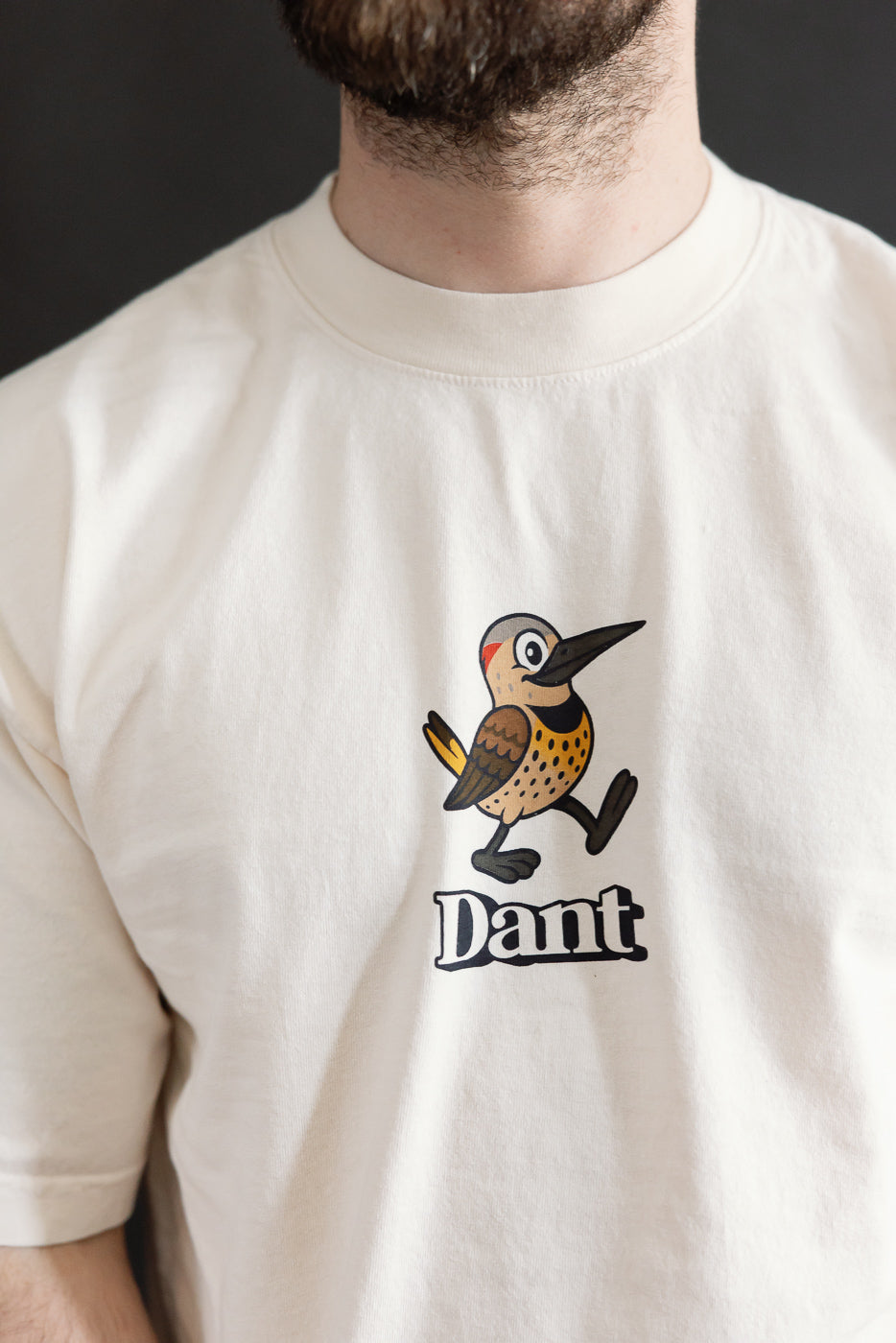 6.5oz - Dant Northern Flicker T-Shirt - Ecru features a cartoon-style Northern Flicker bird striding forward, paired with the Dant logo, showcasing playful design on a light-colored cotton shirt.