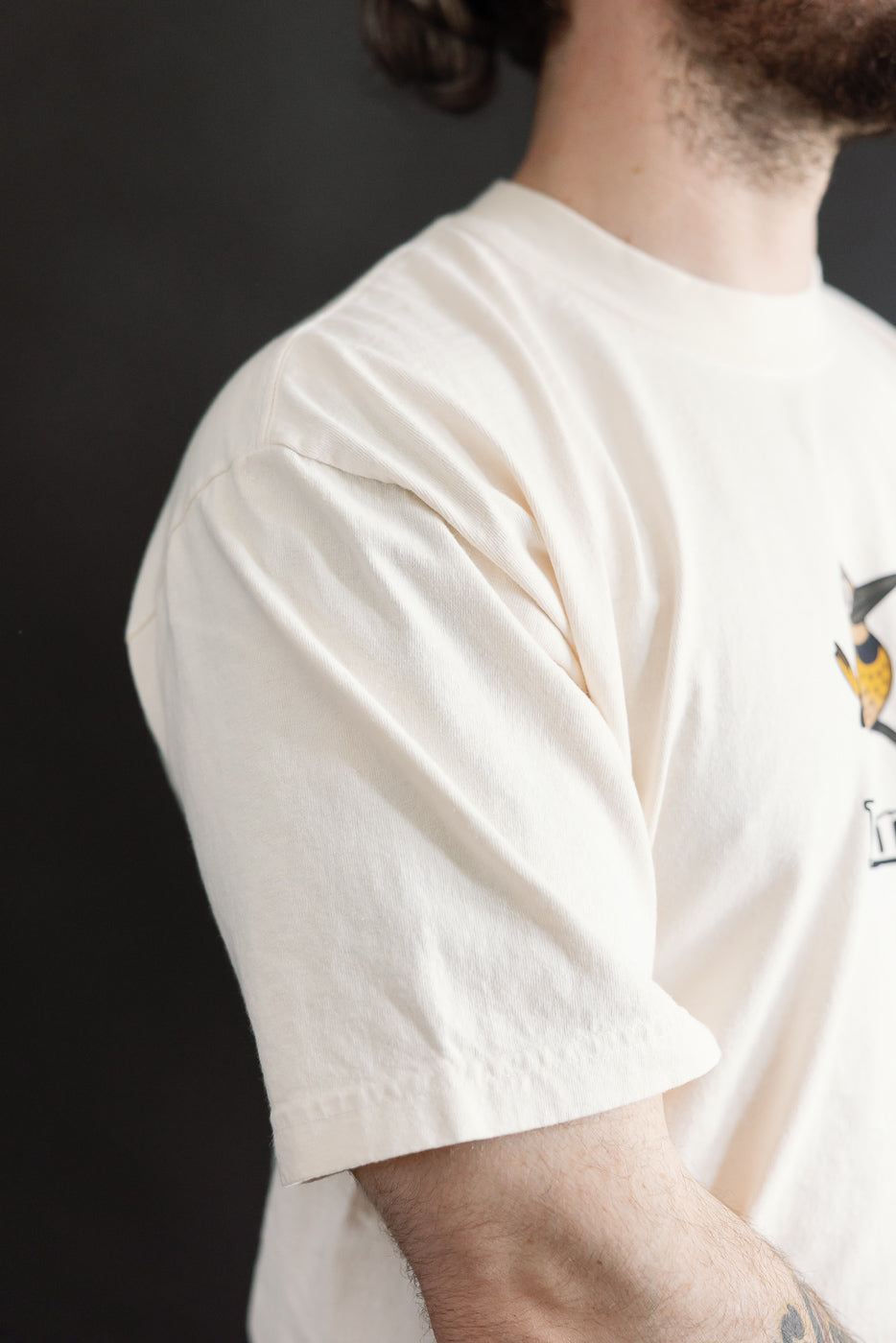 6.5oz - Dant Northern Flicker T-Shirt - Ecru: Close-up of cream cotton tee with yellow and black stylized bird graphic on chest, showcasing the design's intricate detail and casual elegance.