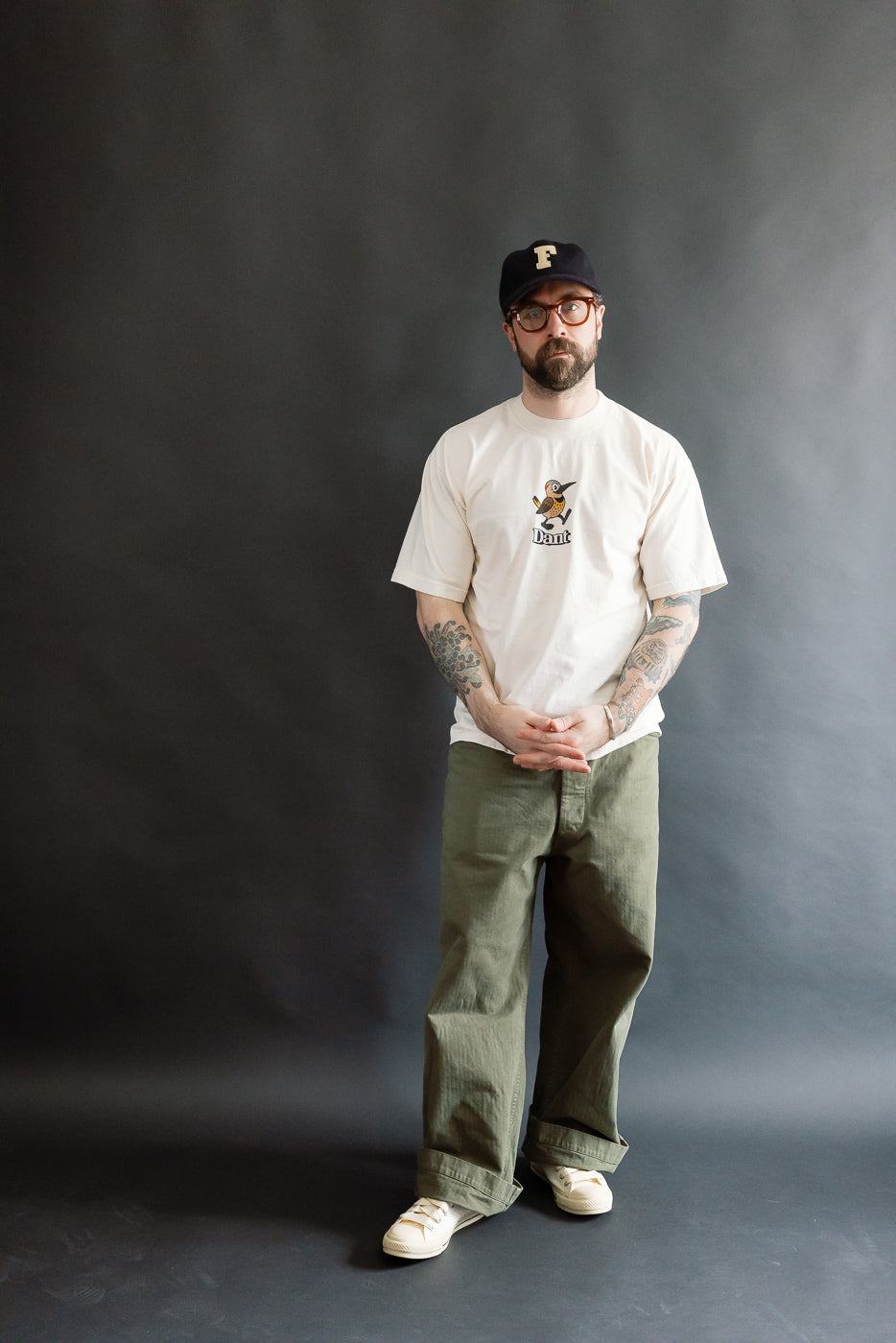 Man wearing the 6.5oz - Dant Northern Flicker T-Shirt - Ecru, showcasing a cartoon bird graphic. Paired with olive trousers and canvas sneakers, highlighting Dant's heritage-inspired style.