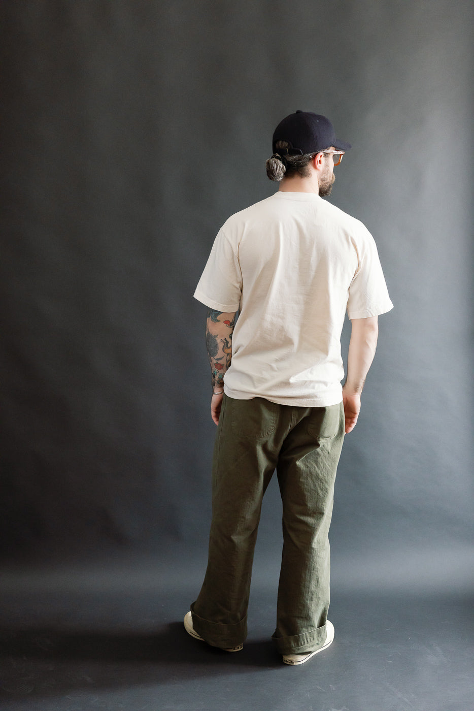 Model showcases the Dant 6.5oz Northern Flicker T-Shirt in Ecru, featuring a relaxed fit, thick ribbed collar, and custom print design, paired with olive-green trousers and canvas sneakers.