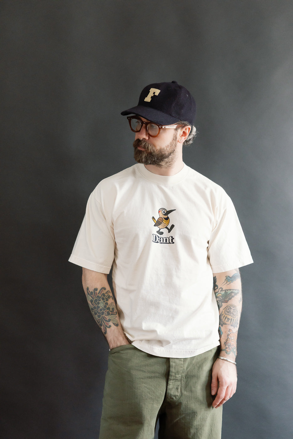 A bearded man models the 6.5oz - Dant Northern Flicker T-Shirt - Ecru, featuring a cartoon woodpecker graphic, styled with olive-green pants and tortoiseshell glasses, highlighting Dant's heritage-inspired aesthetic.