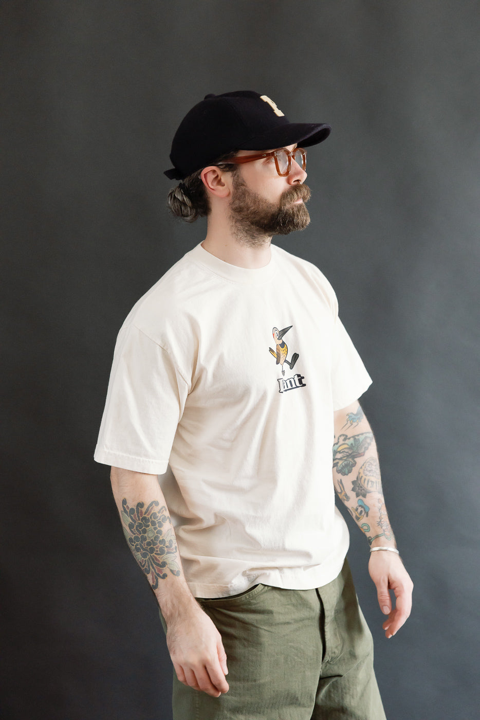 Profile view of a bearded man wearing the 6.5oz - Dant Northern Flicker T-Shirt - Ecru, featuring a stylized bird and mnt logo, paired with olive-green trousers and a black cap.
