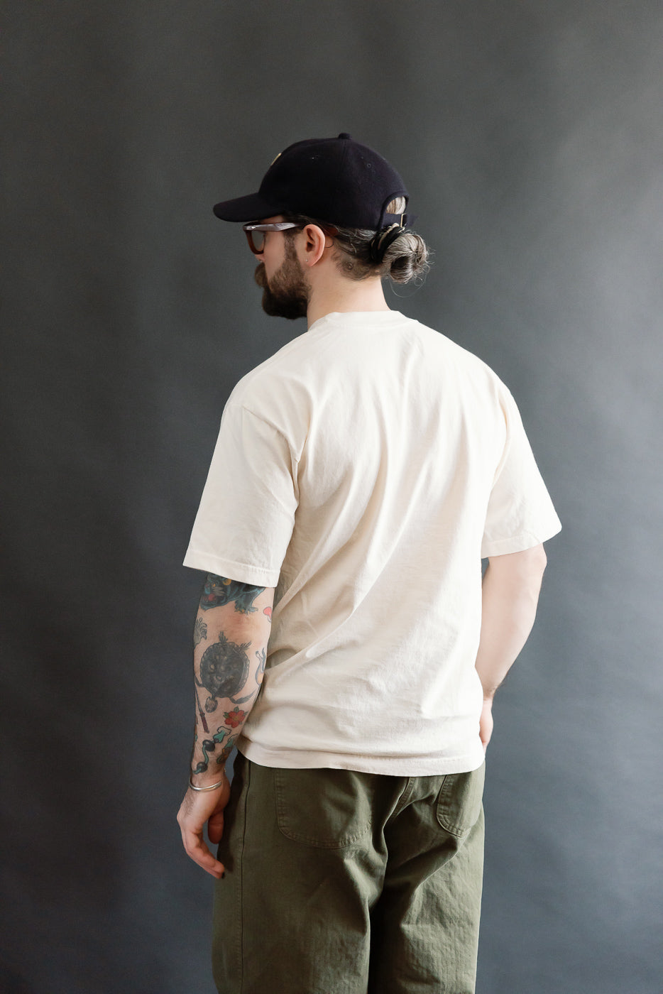 A bearded man models the 6.5oz - Dant Northern Flicker T-Shirt - Ecru, showcasing its oversized fit. He pairs it with olive-green pants and a backward black cap, embodying a casual, modern look.