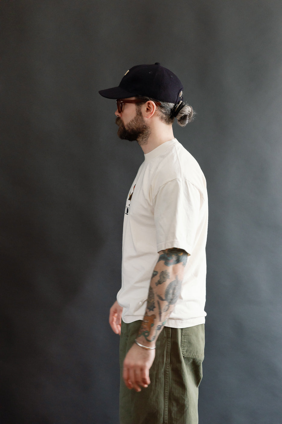 A bearded man models the 6.5oz - Dant Northern Flicker T-Shirt, showcasing its oversized fit and thick ribbed collar, styled with olive-green pants and a black cap.