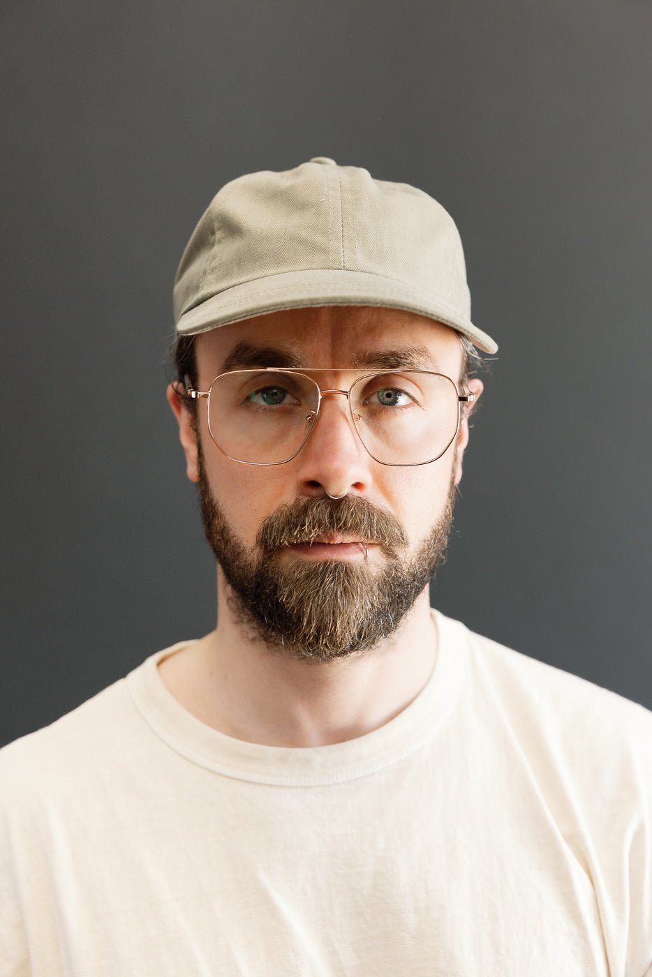 Man wearing the Dry Wax Cotton Baseball Hat - Sage, showcasing its mid-profile crown and flat brim. The cap's earthy tone complements his cream T-shirt, emphasizing the hat's vintage, utilitarian style.