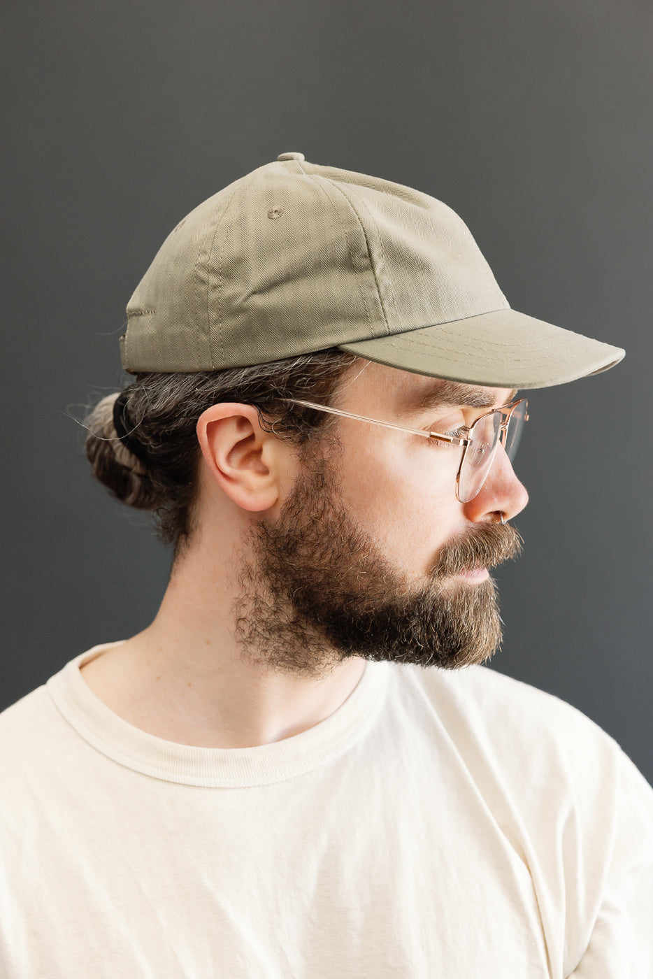 Side-profile of a man wearing Dehen’s Dry Wax Cotton Baseball Hat - Sage, highlighting its unstructured six-panel design and clear-framed eyeglasses. The hat features a flat brim and self-fabric adjustable strap.