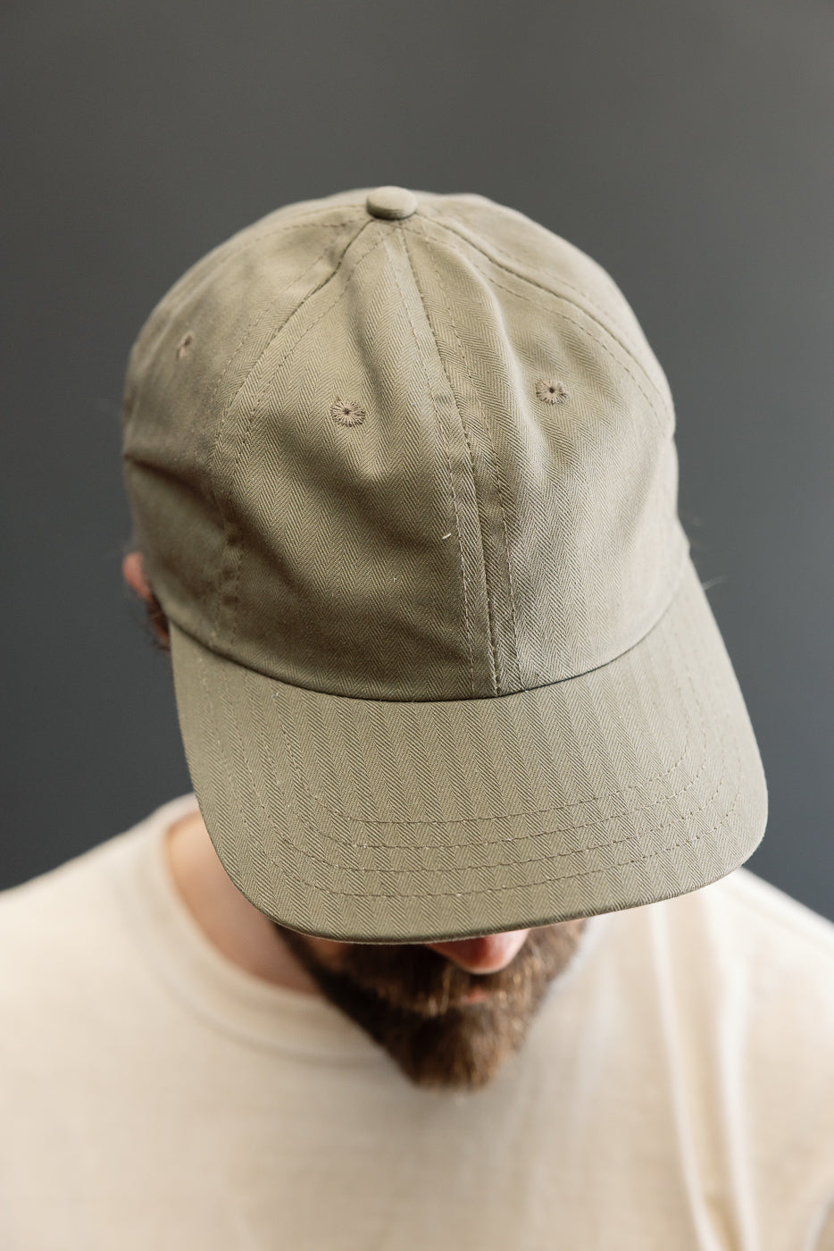 Close-up of the Dry Wax Cotton Baseball Hat - Sage, showcasing its six-panel construction, herringbone-textured fabric, and antique brass slider. The cap is worn low, partially obscuring the wearer's face.