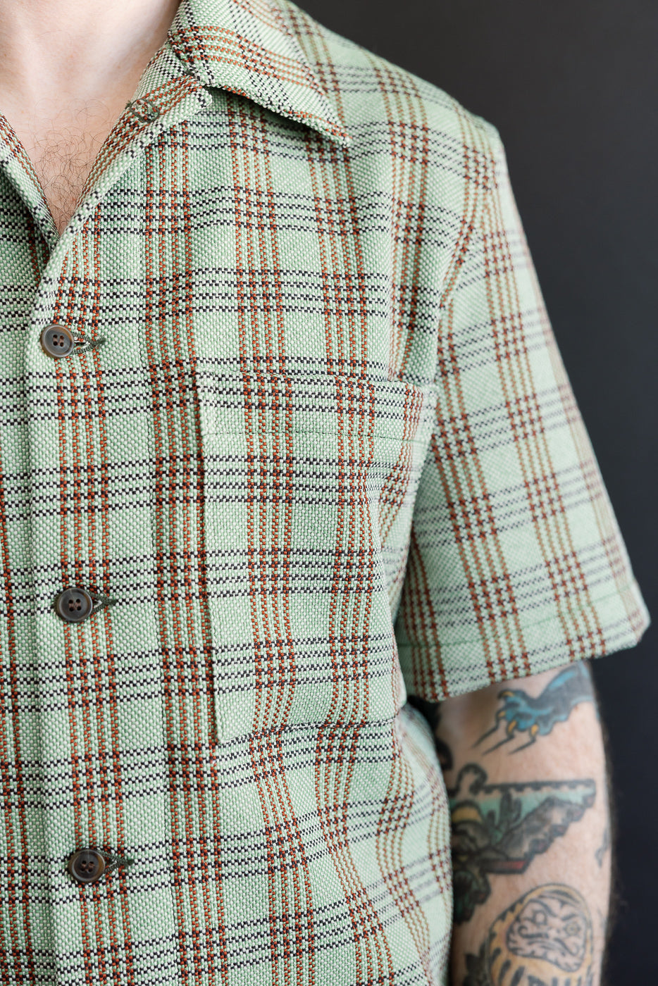 Close-up front view of green plaid lounge shirt on male model, showing buttons, collar, chest pocket, and fabric texture. Model is 5'9