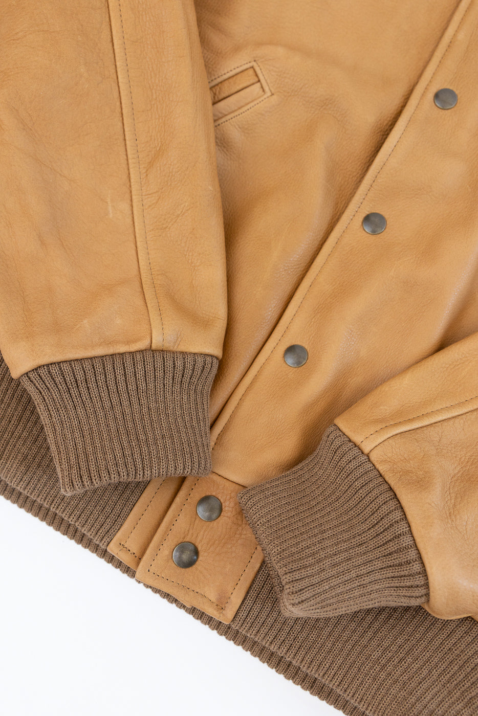 Close-up of front of tan leather jacket showing snap buttons, ribbed cuffs, and pocket detail.