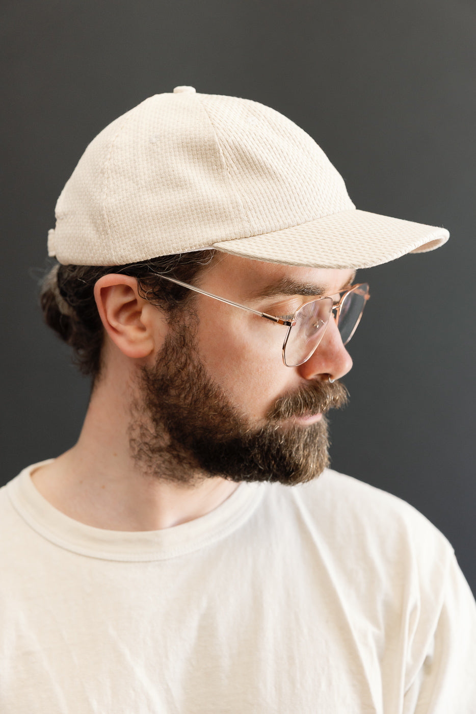 Sashiko Cotton Baseball Hat - Natural: Close-up of a man wearing a cream-colored, unstructured baseball cap made from natural sashiko cotton, featuring a 6-panel crown, pre-curved brim, and brass-buckled leather strap.