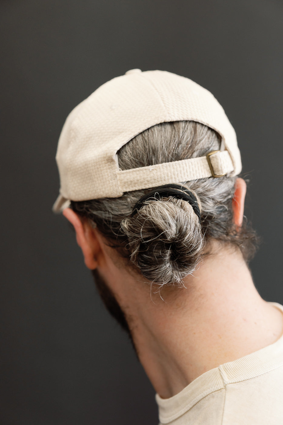 Person wearing the Sashiko Cotton Baseball Hat - Natural, featuring an unstructured 6-panel crown and pre-curved brim, with salt-and-pepper hair in a bun, against a plain, dark background.