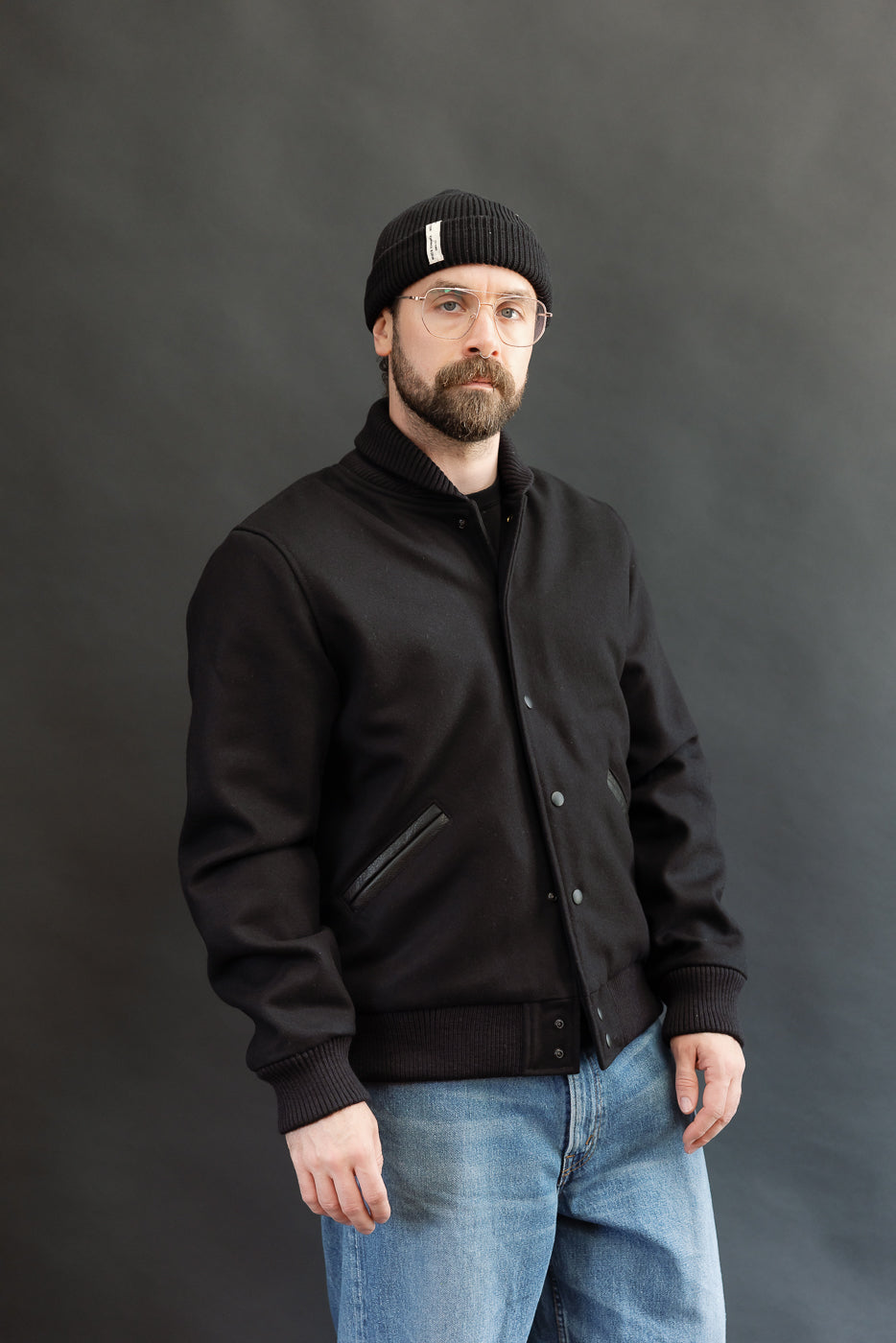 male model wearing an all black varsity jacket with black beanie, washed jeans, 3/4, front view