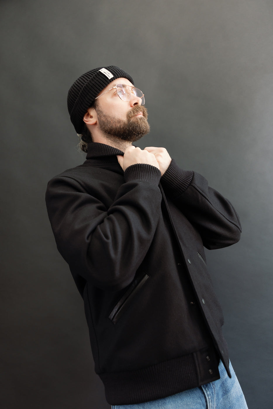 male model wearing an all black varsity jacket with black beanie, washed jeans, 3/4, front view
