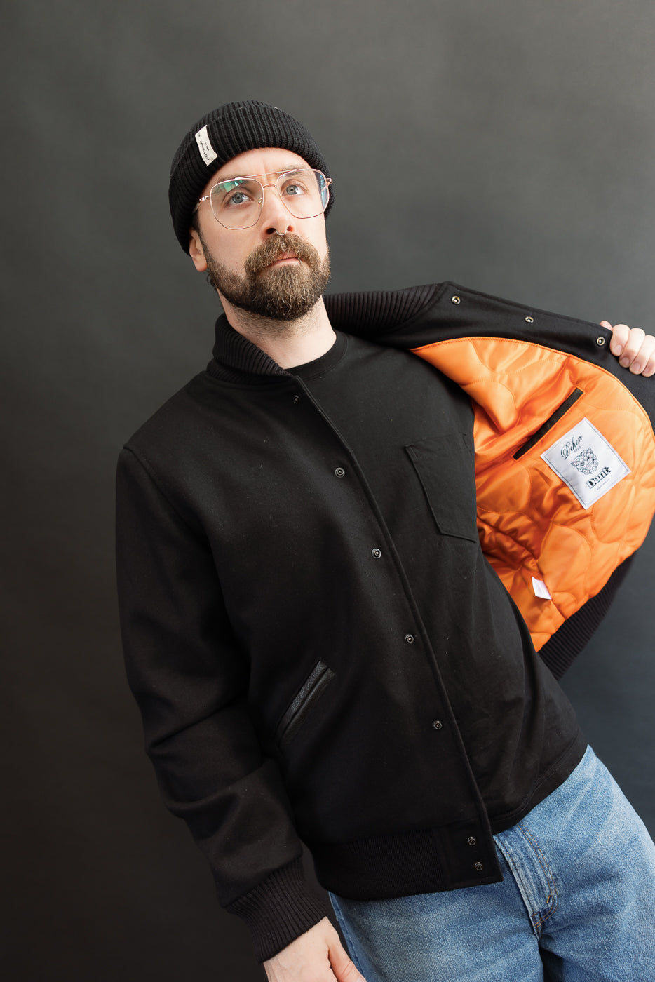 male model wearing an all black varsity jacket with black beanie, washed jeans, opening jacket to show liner, 3/4 body, front view