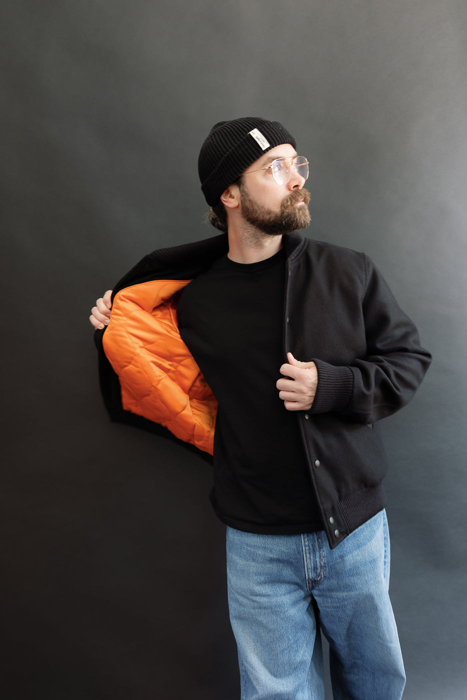 male model wearing an all black varsity jacket with black beanie, washed jeans, opening jacket to show liner, 3/4 body, front view
