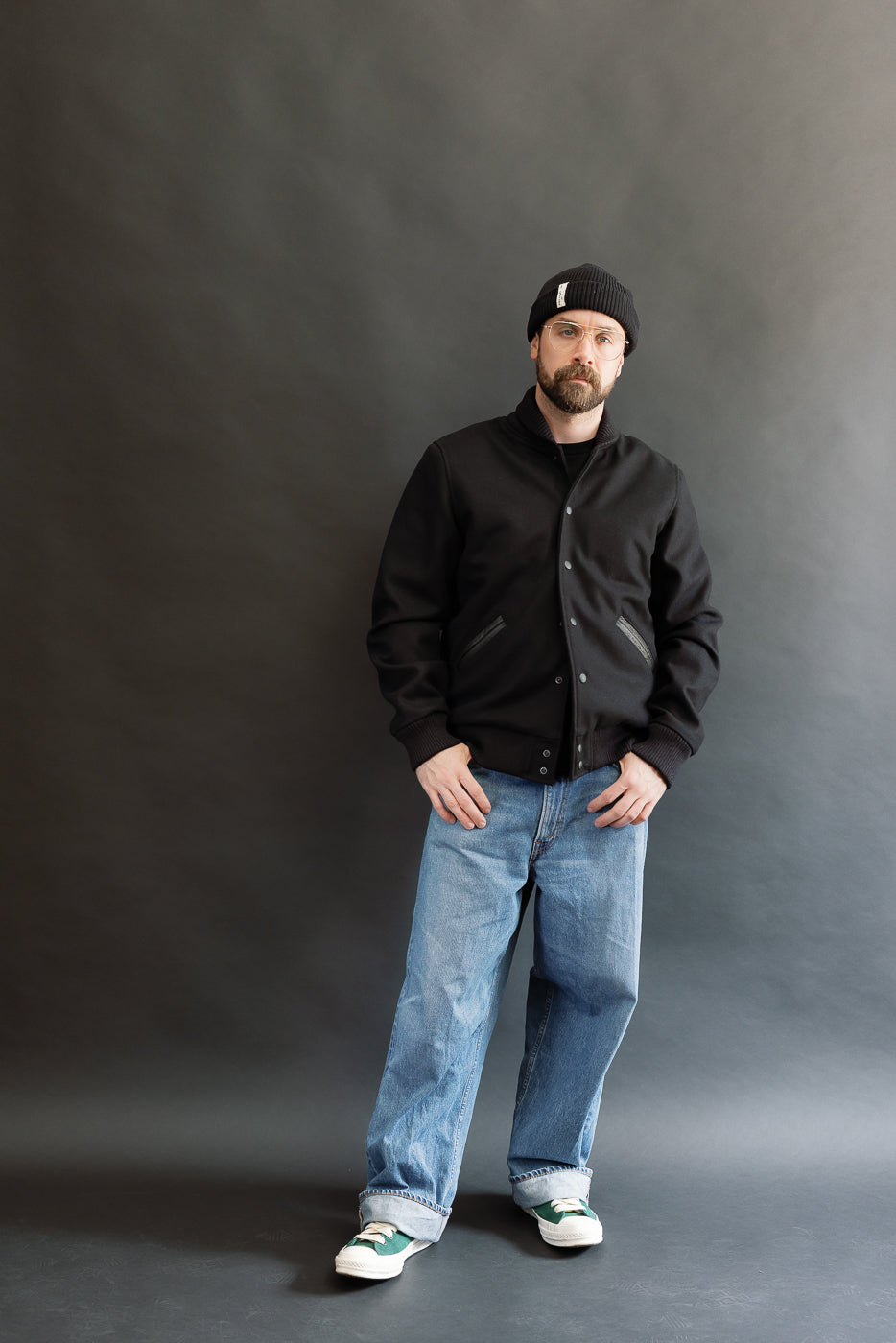 male model wearing an all black varsity jacket with black beanie, washed jeans, and green chucks, full body, front view