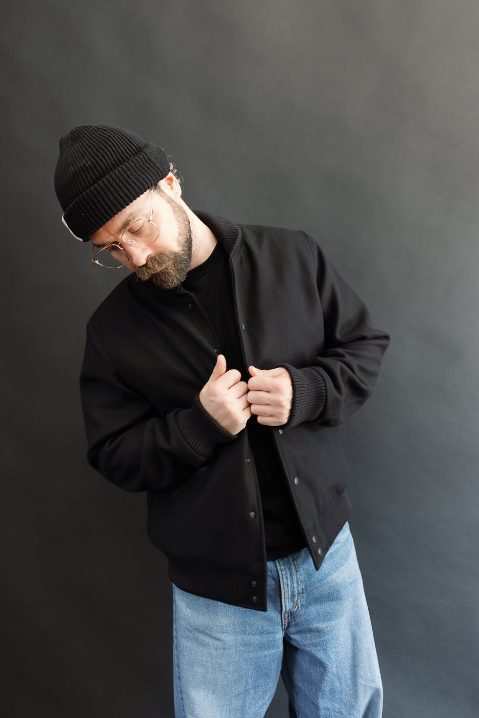 male model wearing an all black varsity jacket with black beanie, washed jeans, 3/4 body, front view