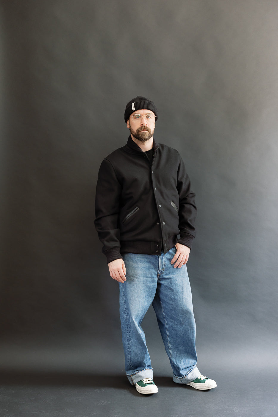 male model wearing an all black varsity jacket with black beanie, washed jeans, and green chucks, full body, front view