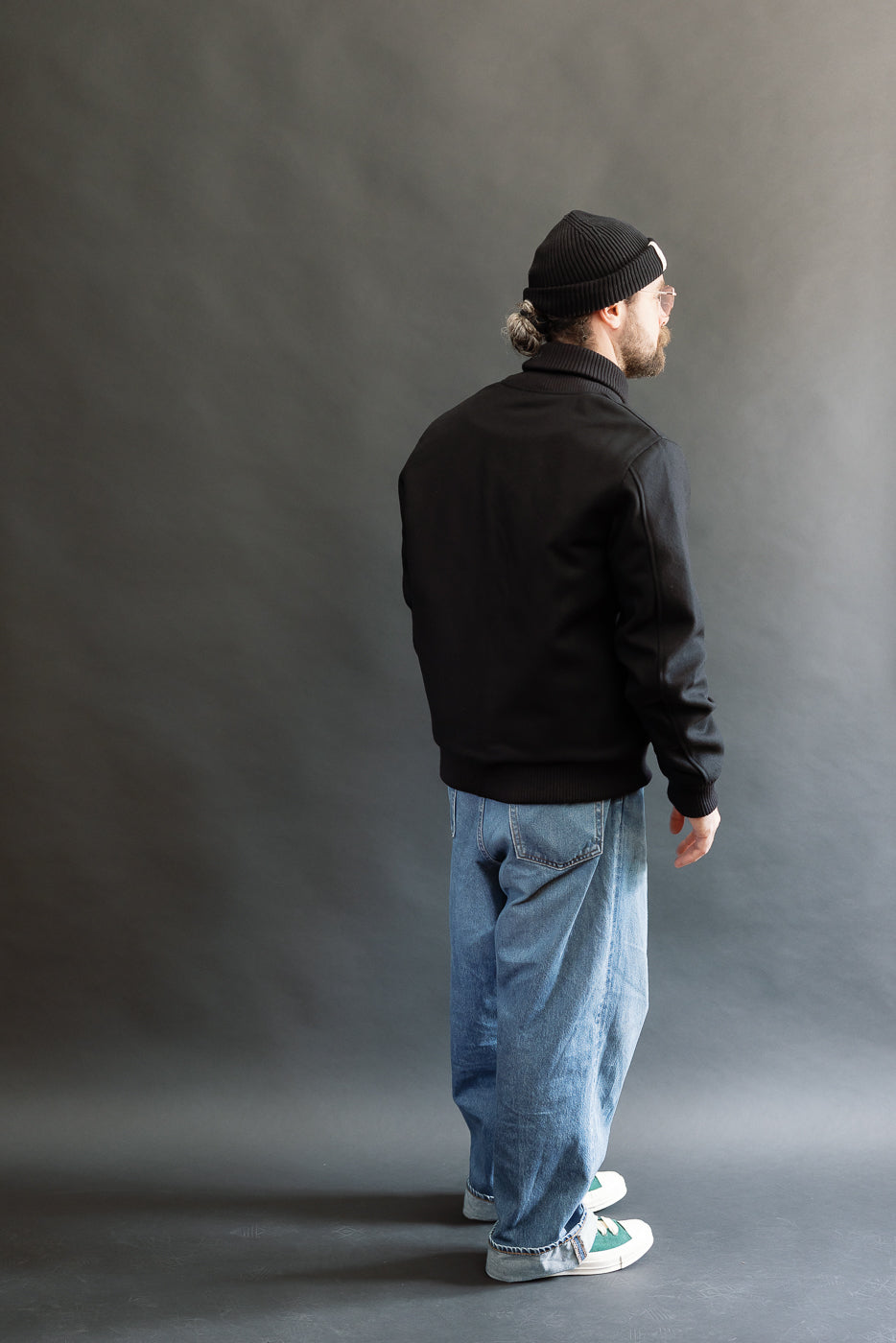male model wearing an all black varsity jacket with black beanie, washed jeans, and green chucks, full body, back view