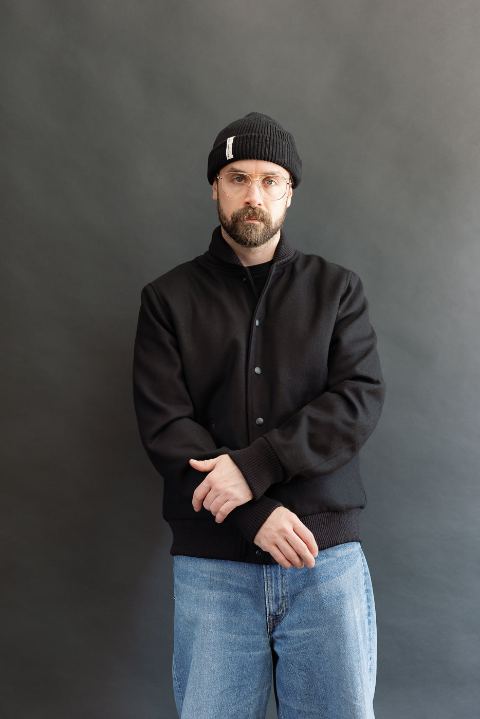 male model wearing an all black varsity jacket with black beanie, washed jeans, 3/4, front view
