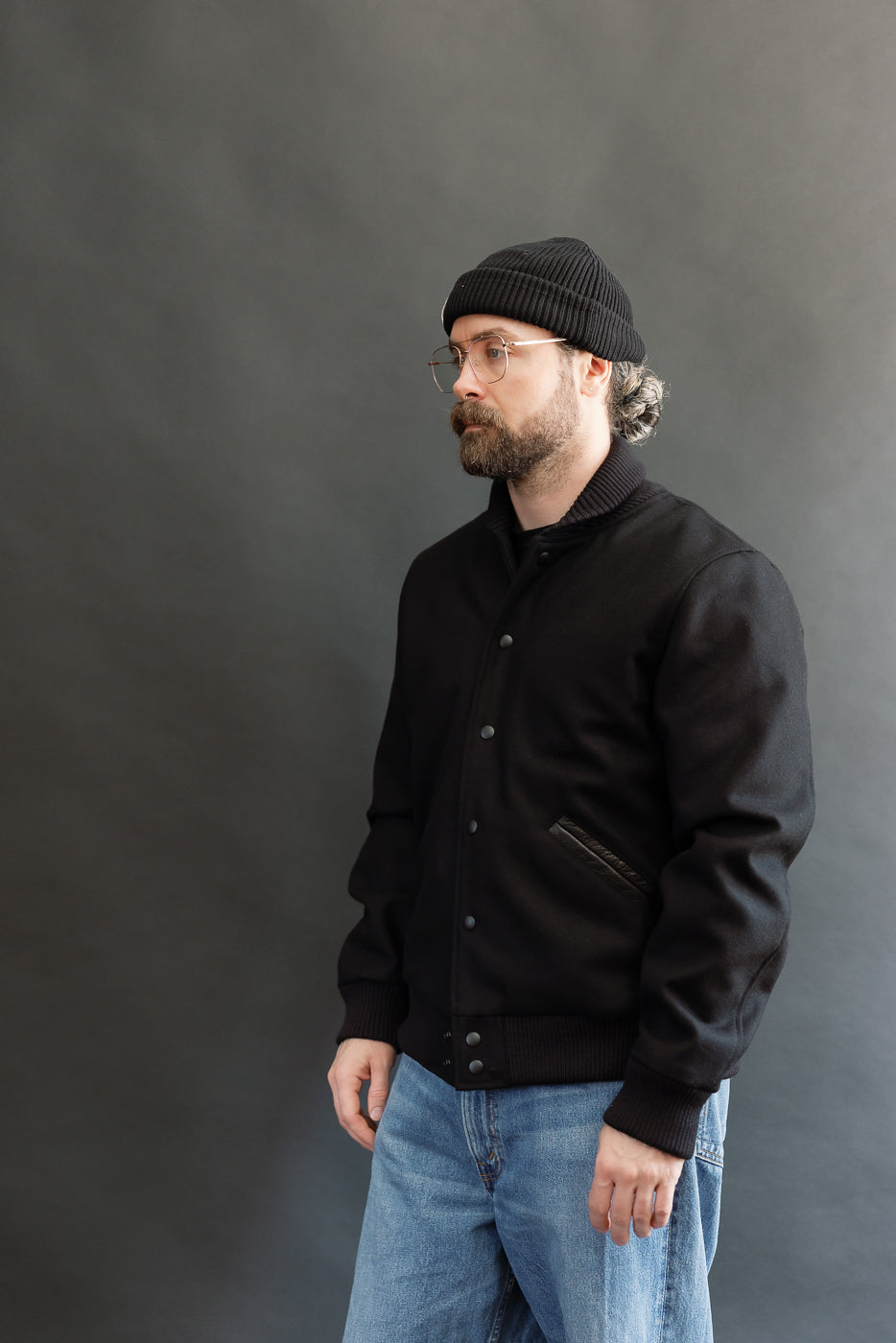 male model wearing an all black varsity jacket with black beanie, washed jeans, 3/4, side view