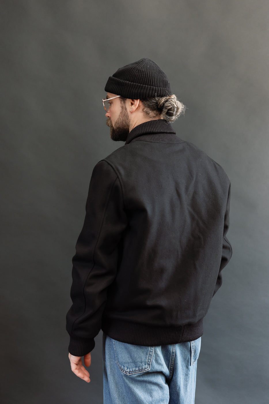 male model wearing an all black varsity jacket with black beanie, washed jeans, 3/4, back view