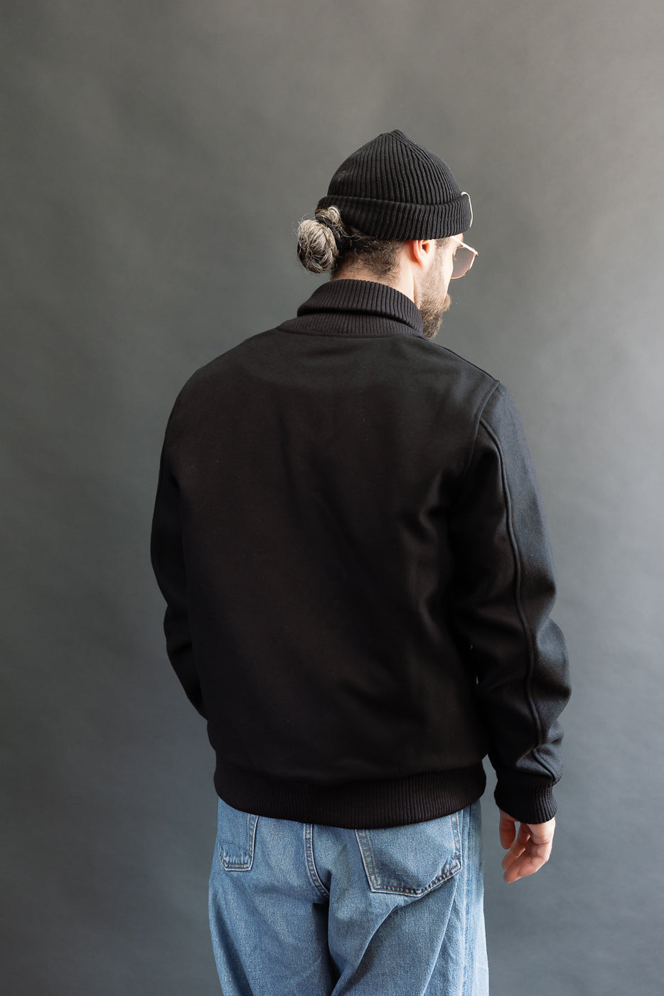 male model wearing an all black varsity jacket with black beanie, washed jeans, 3/4, back view