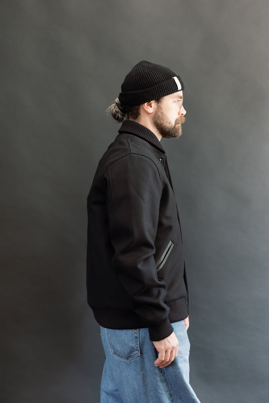 male model wearing an all black varsity jacket with black beanie, washed jeans, 3/4, side view