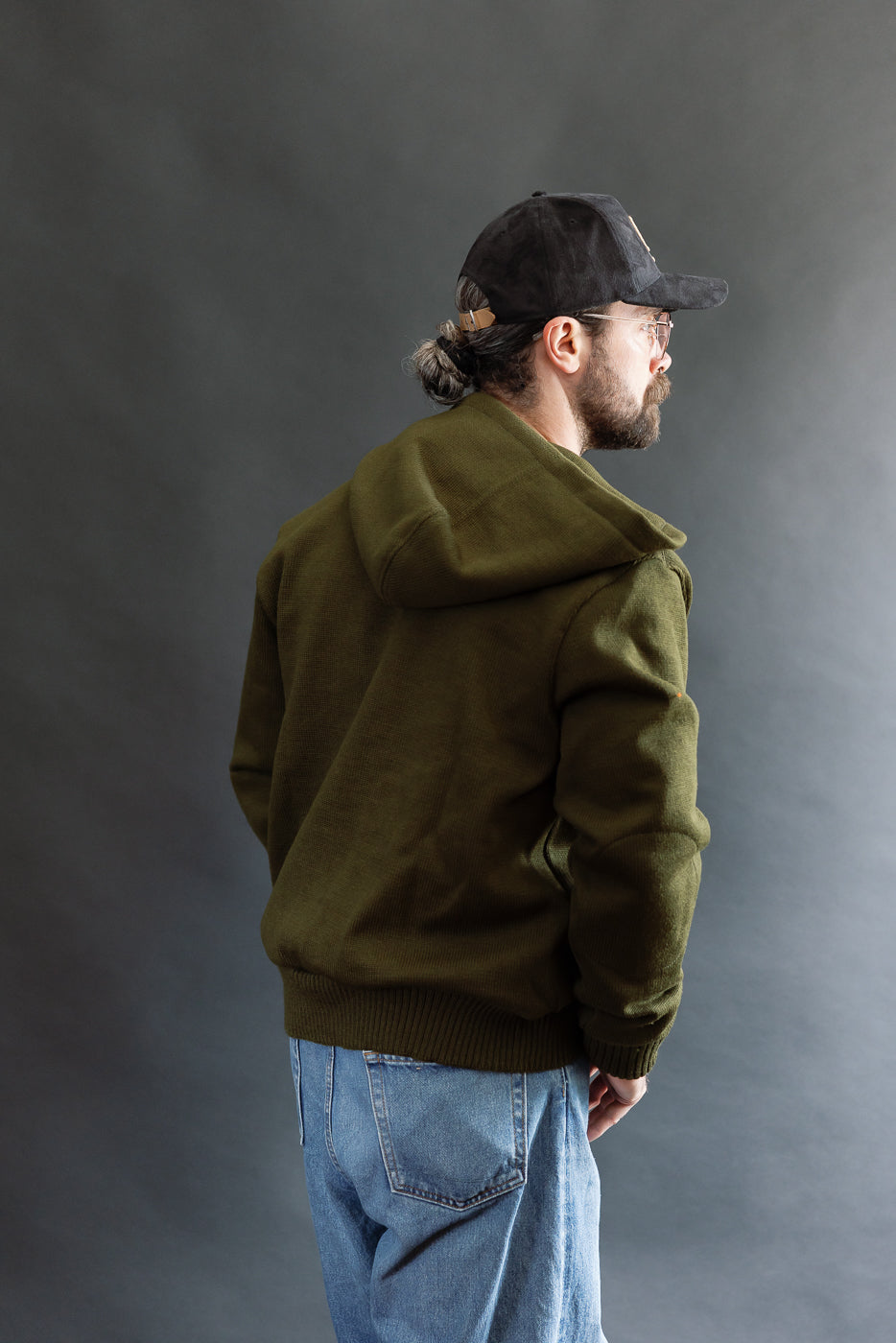 male model wearing an open green knitted zip up hoodie with a black hat, t-shirt, and washed jeans, 3/4 body, back view