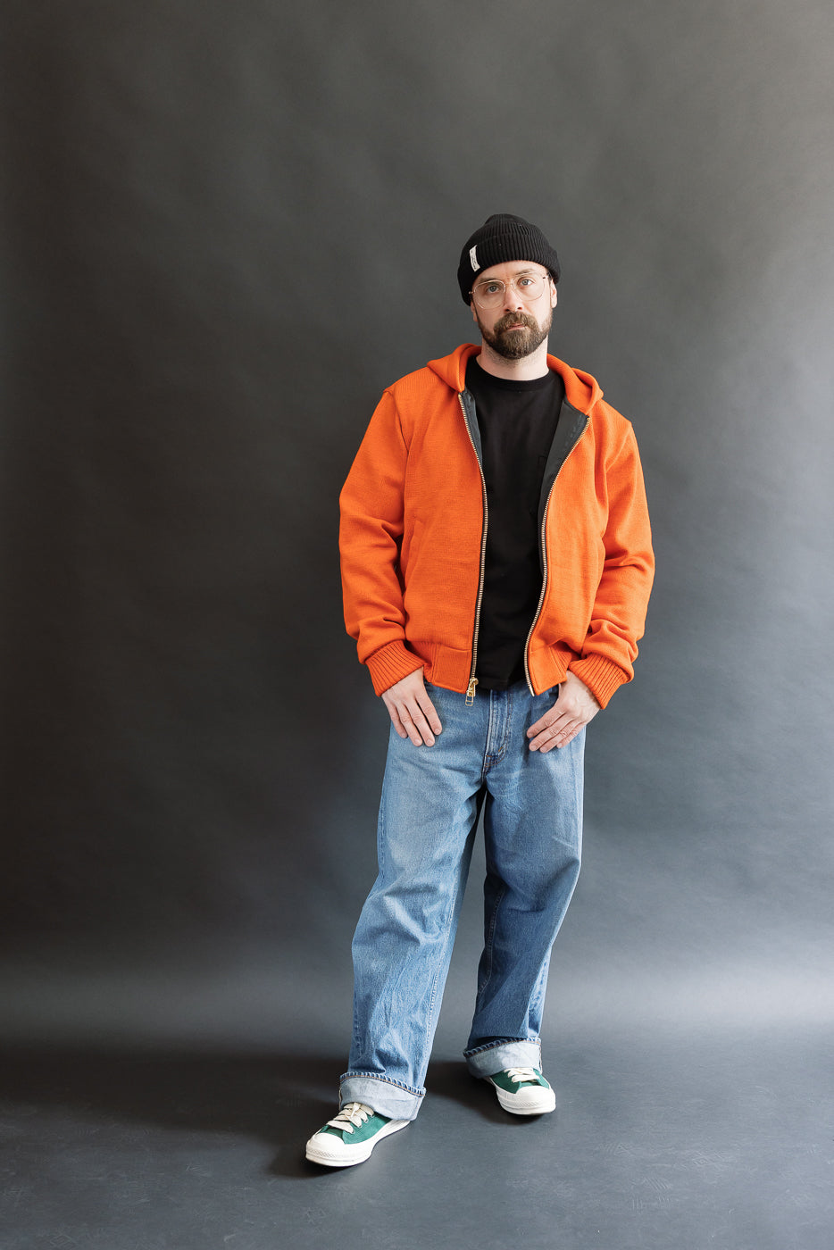 Male model wearing a knitted orange hoodie (open), black beanie, blackshirt, and washed blue jeans standing against a gray background, full body, front view