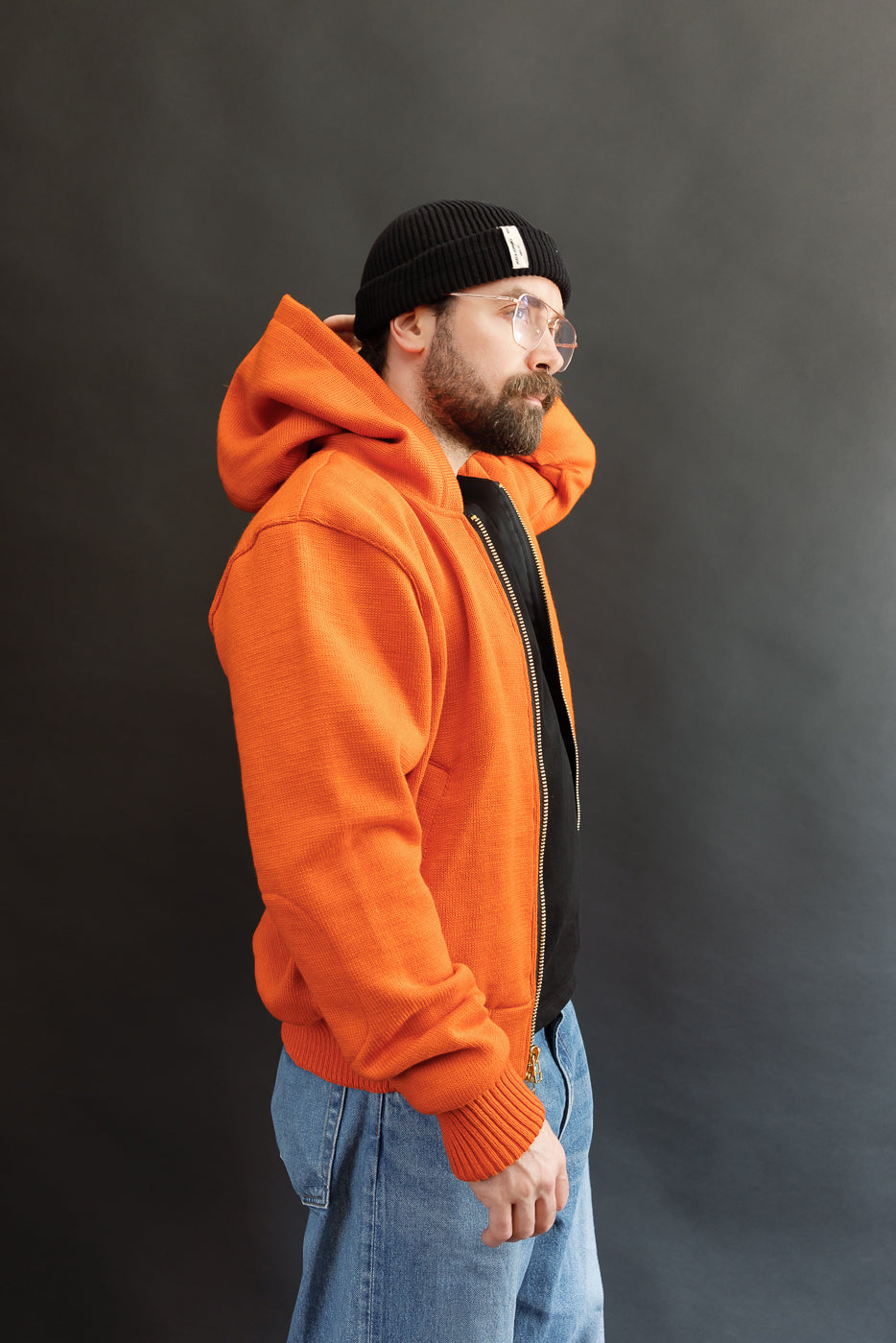 Male model wearing a knitted orange hoodie (open), black beanie, blackshirt, and washed blue jeans standing against a gray background, 3/4 body, side view
