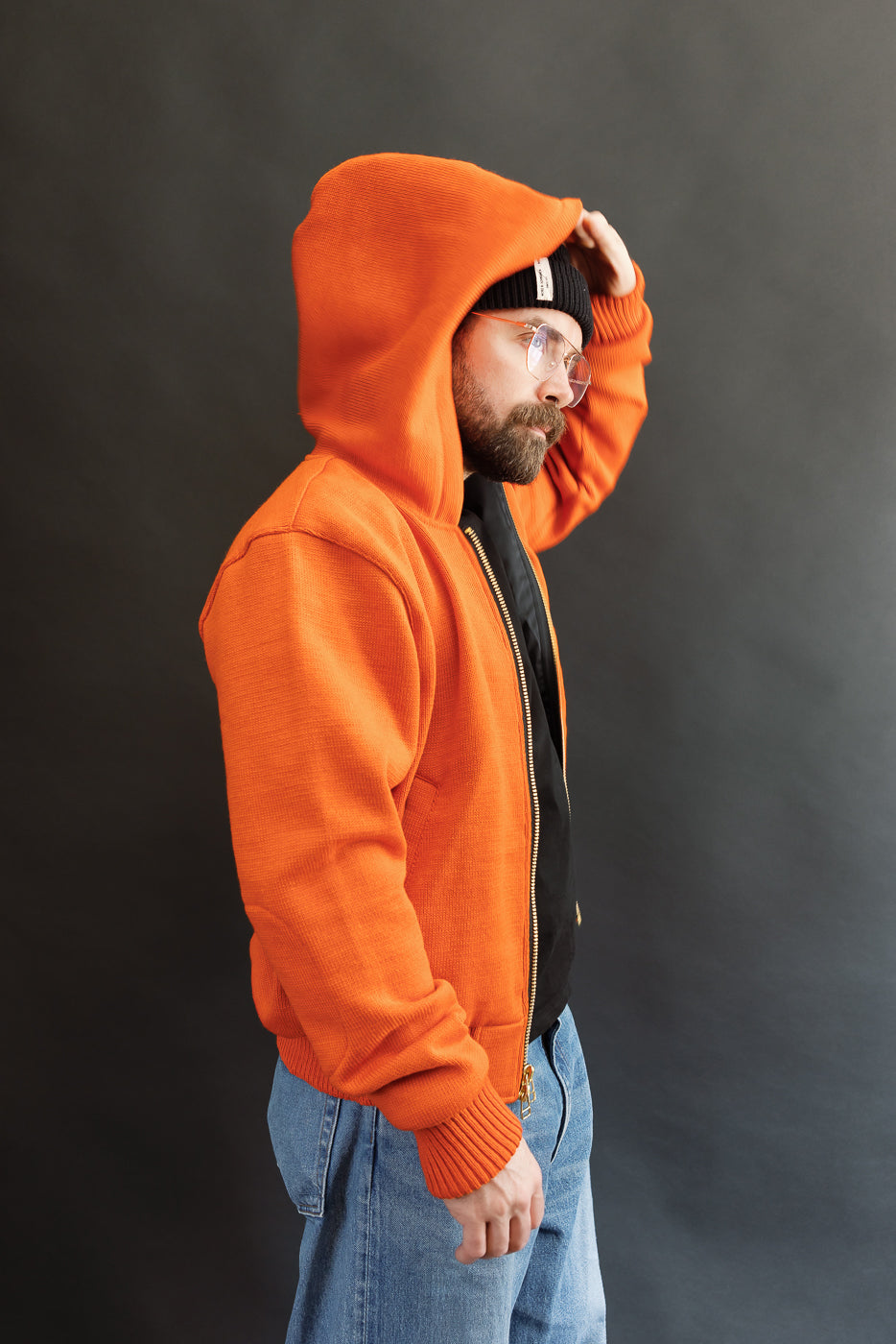 Male model wearing a knitted orange hoodie (open), black beanie, blackshirt, and washed blue jeans standing against a gray background, 3/4 body, side view, hood up