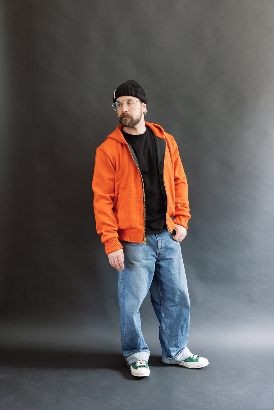 Male model wearing a knitted orange hoodie (open), black beanie, blackshirt, and washed blue jeans standing against a gray background, full body, front view