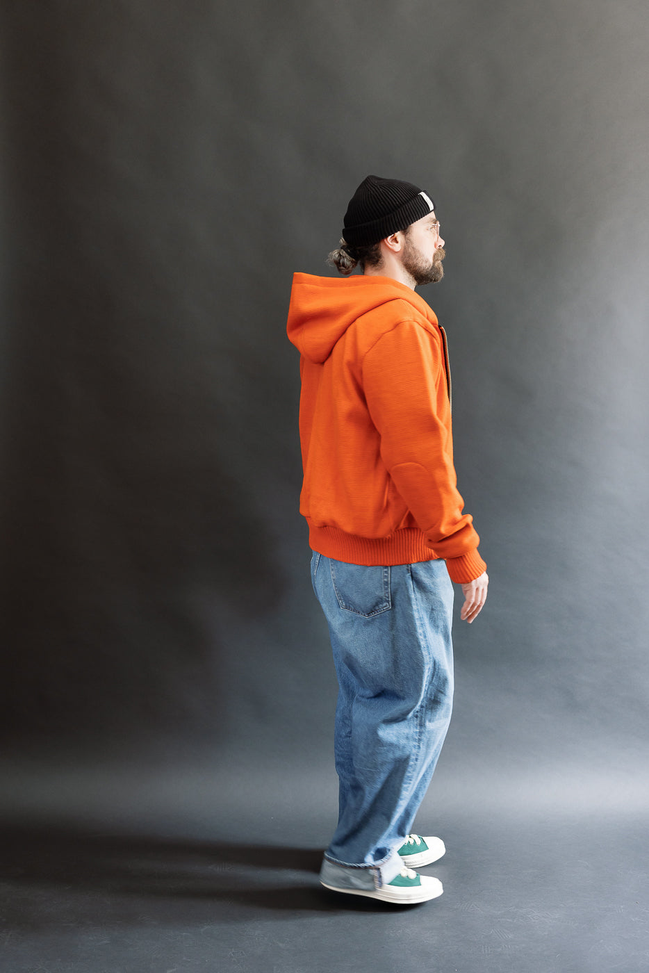 Male model wearing a knitted orange hoodie (open), black beanie, blackshirt, and washed blue jeans standing against a gray background, full body, side view