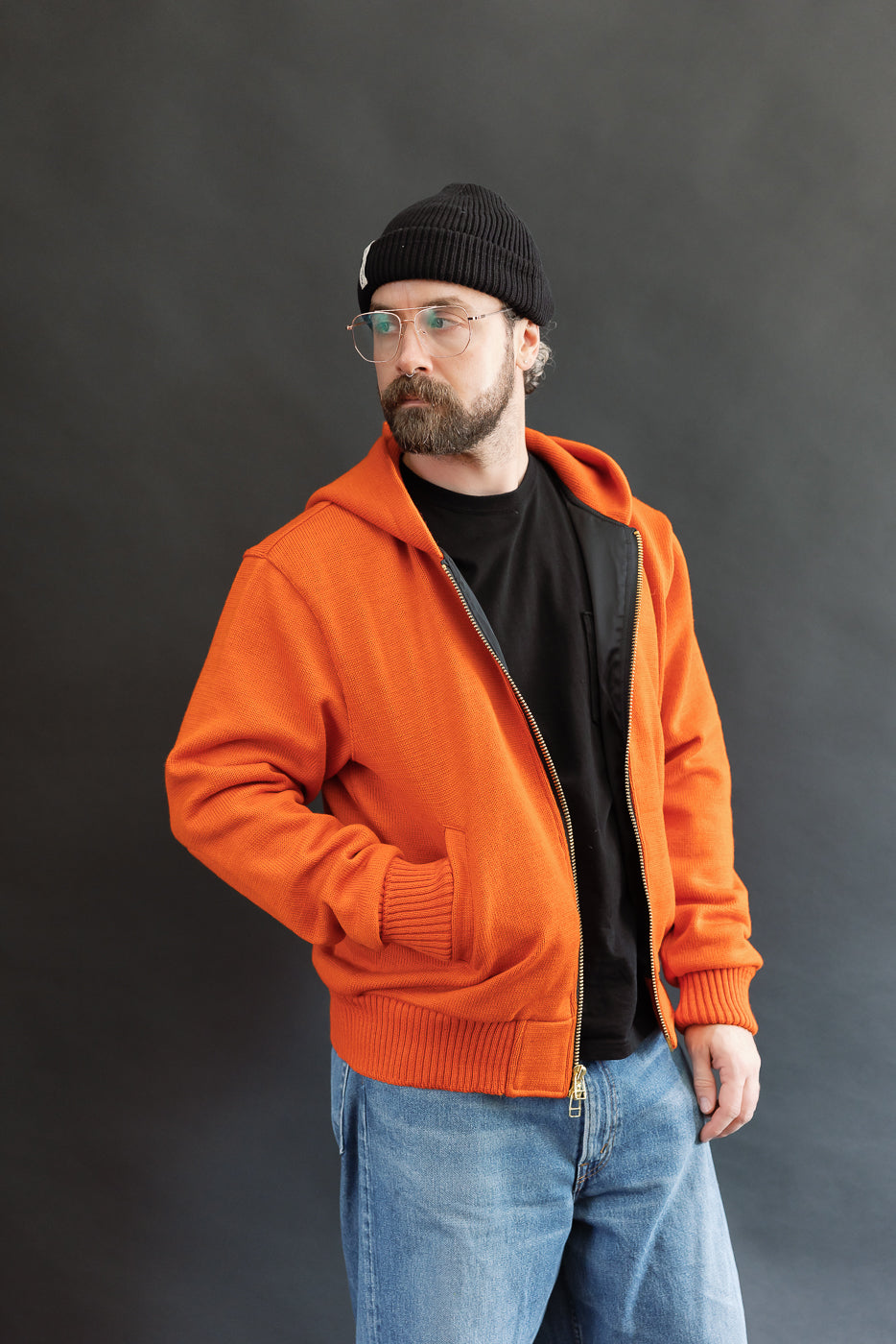 Male model wearing a knitted orange hoodie (open), black beanie, blackshirt, and washed blue jeans standing against a gray background, 3/4 body, front view