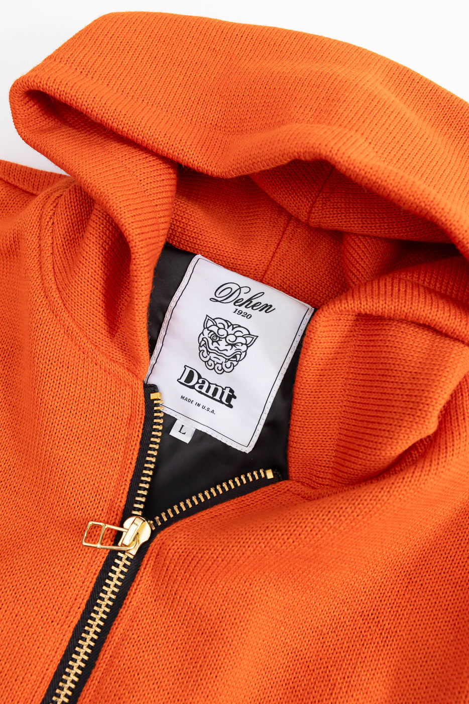 Close-up of an orange hoodie with a focus on a white dehen x dant logo brand label on a white background