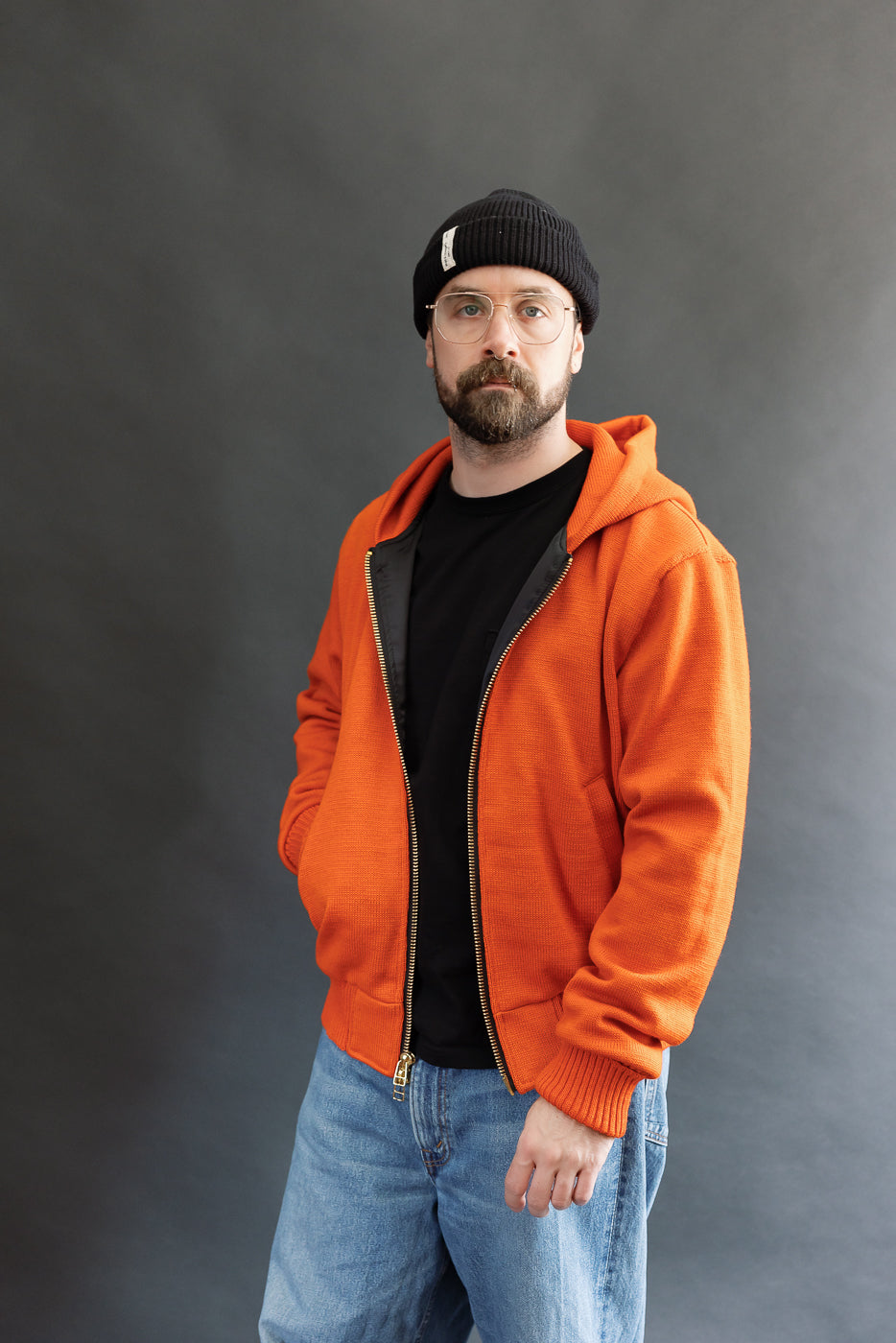Male model wearing a knitted orange hoodie (open), black beanie, blackshirt, and washed blue jeans standing against a gray background, 3/4 body, side view