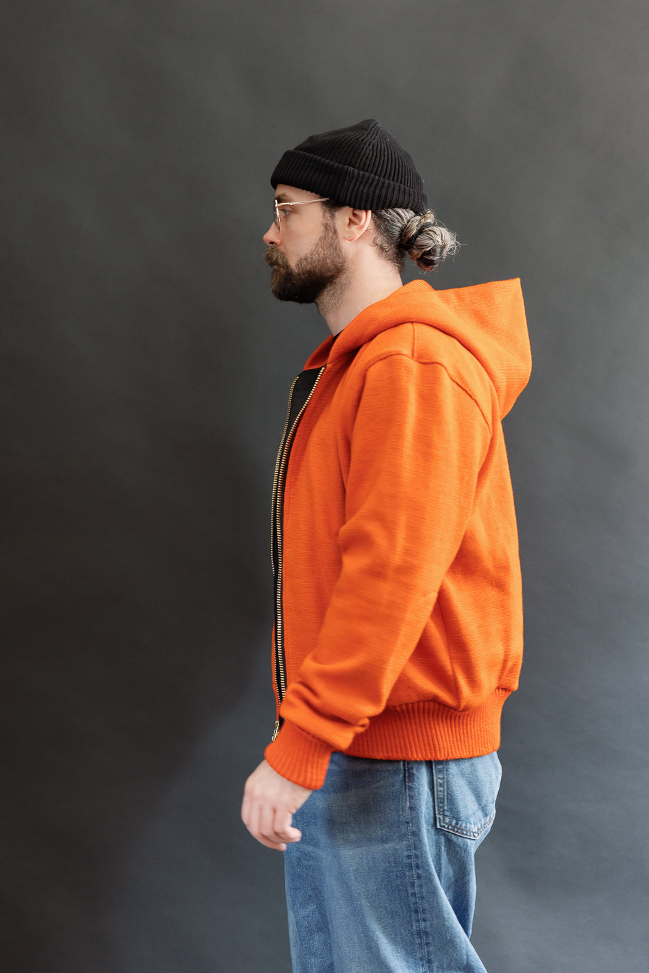 Male model wearing a knitted orange hoodie (open), black beanie, blackshirt, and washed blue jeans standing against a gray background, 3/4 body, side view
