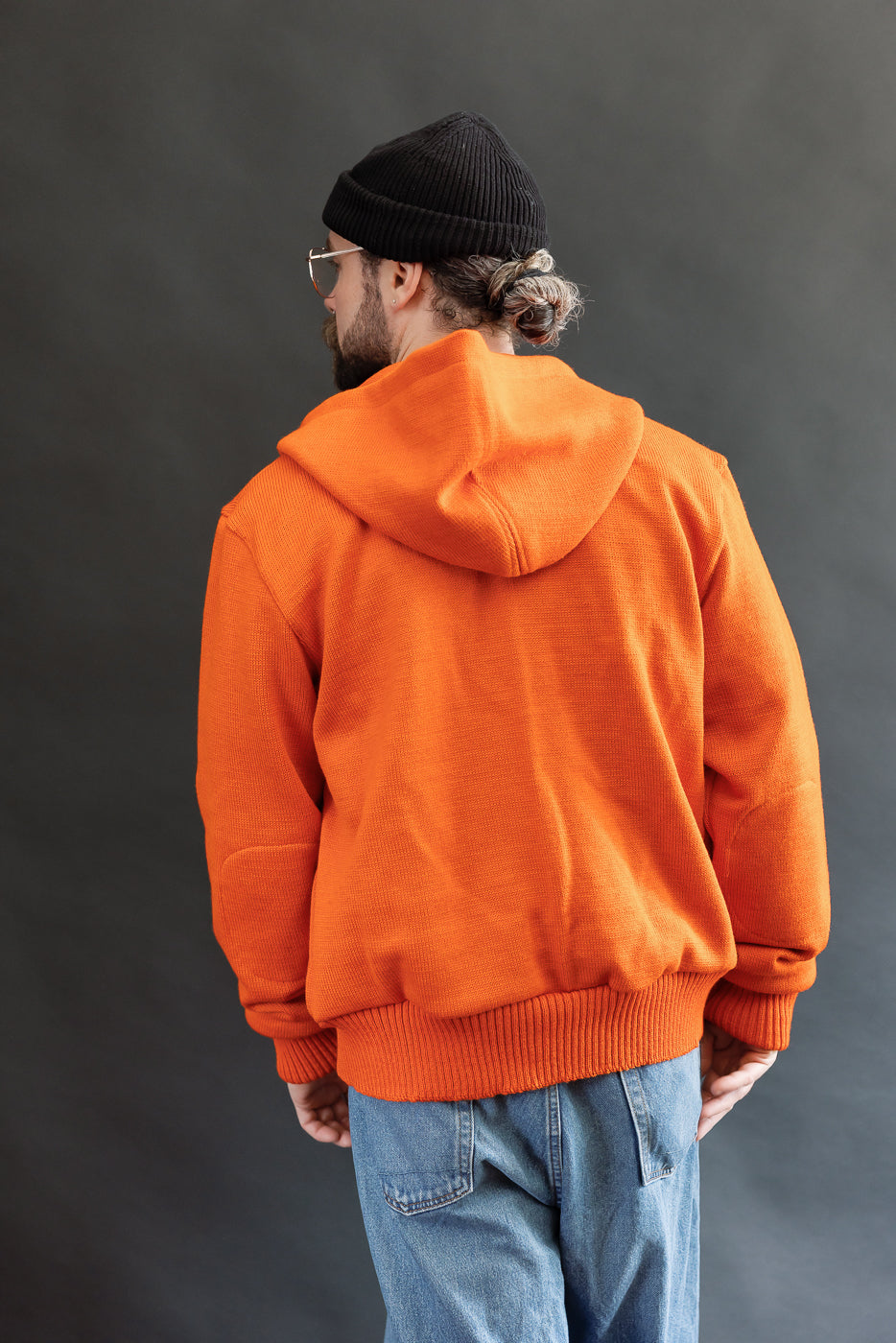 Male model wearing a knitted orange hoodie (open), black beanie, blackshirt, and washed blue jeans standing against a gray background, 3/4 body, back view