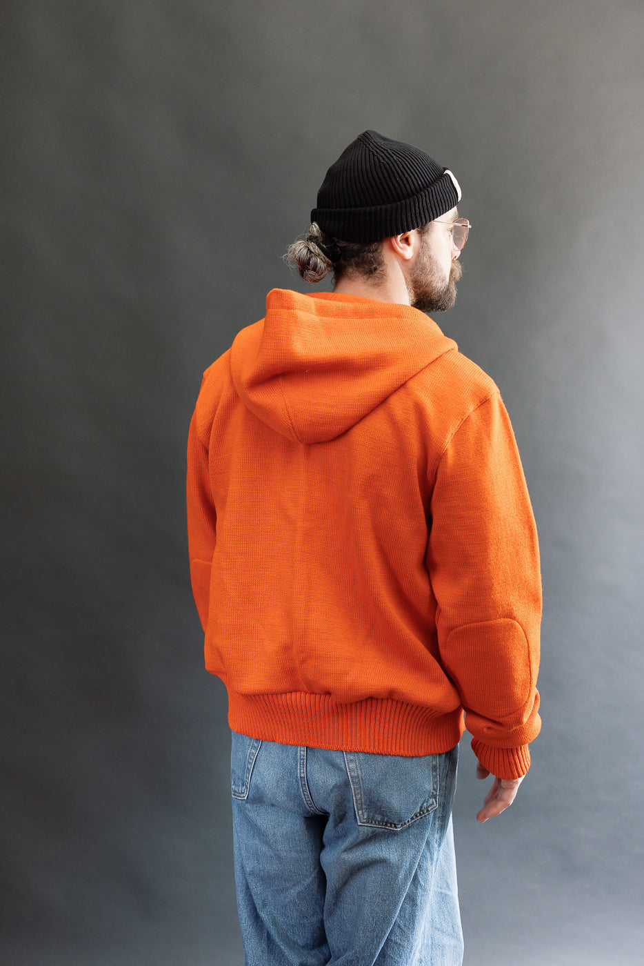 Male model wearing a knitted orange hoodie (open), black beanie, blackshirt, and washed blue jeans standing against a gray background, 3/4 body, back view