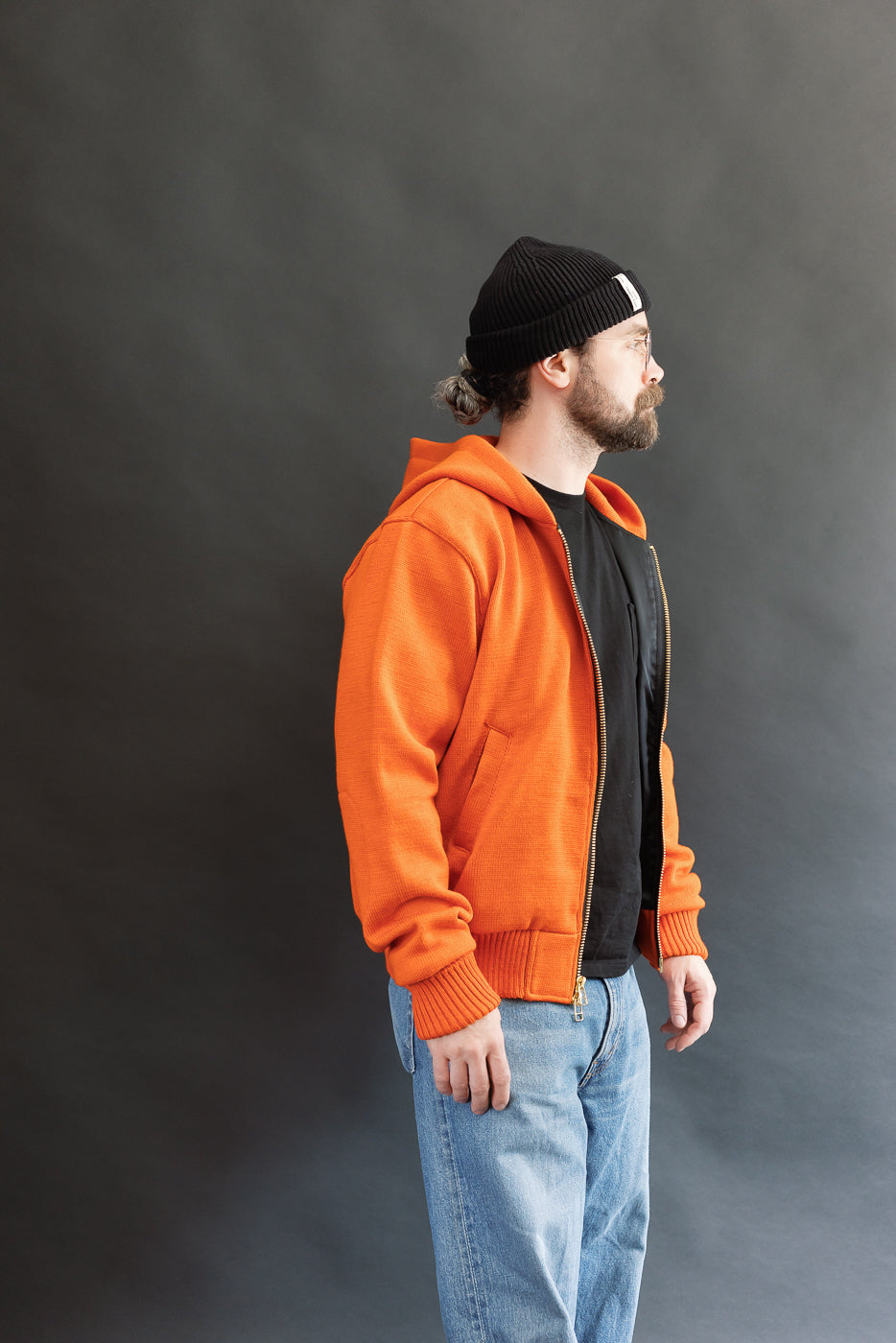 Male model wearing a knitted orange hoodie (open), black beanie, blackshirt, and washed blue jeans standing against a gray background, 3/4 body, side view
